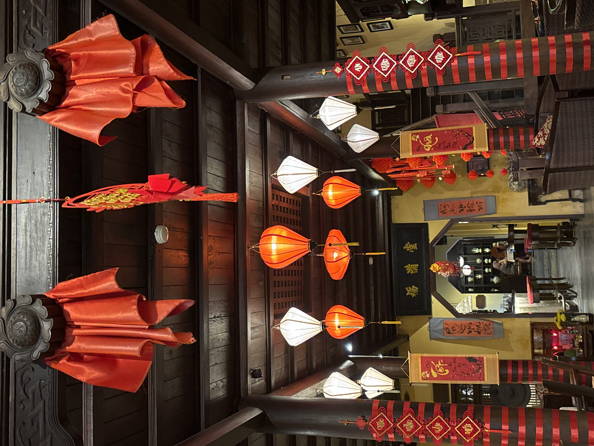 The image shows the interior of a building in Hội An Ward, Vietnam. The ceiling is adorned with dark wooden beams and numerous lanterns. There are three rows of lanterns: the top and bottom rows consist of white lanterns, while the middle row is composed of orange lanterns. The wooden beams are decorated with red banners and what appear to be decorative knots. The architectural details include carved dark wood and ornate decorative elements. In the background, there's a glimpse into another room with tables, chairs, and shelves displaying decorative items. Several people are seated at tables in the background. The overall atmosphere suggests a traditional, cultural, and dimly lit setting.