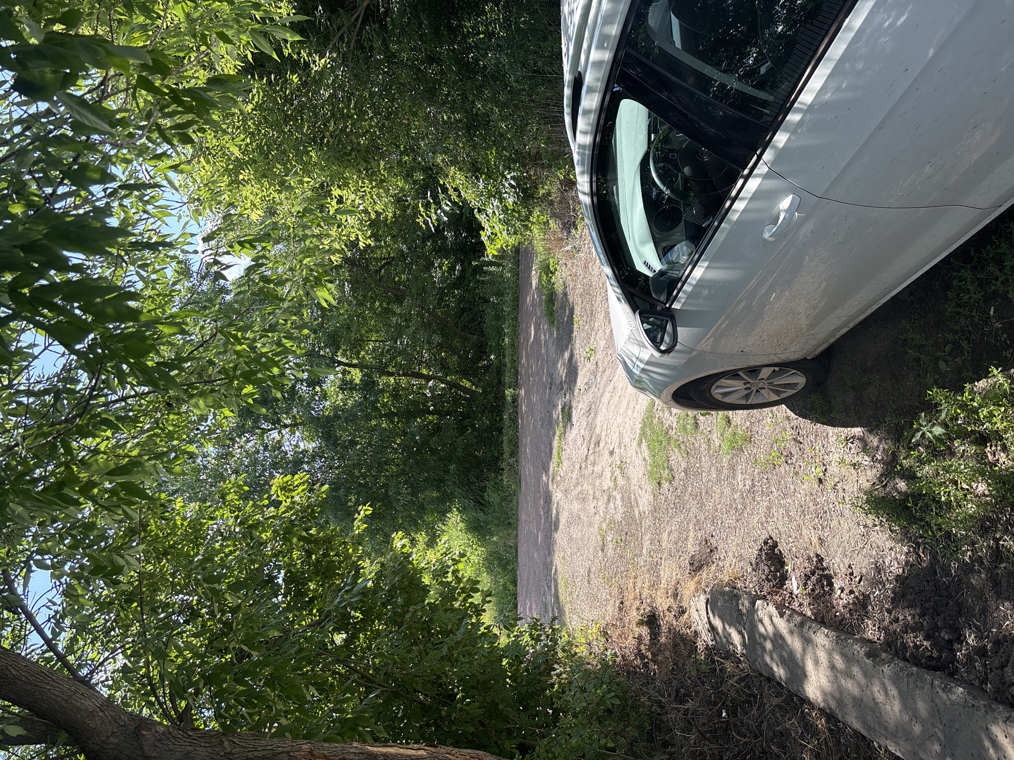 A white car is parked on a dirt road surrounded by trees and greenery. The car is a hatchback, with its sunroof open. The road appears to be unpaved and leads into a wooded area. The sky is blue with some clouds. The image was taken on a sunny day. The location is Pereiaslavske, Ukraine.