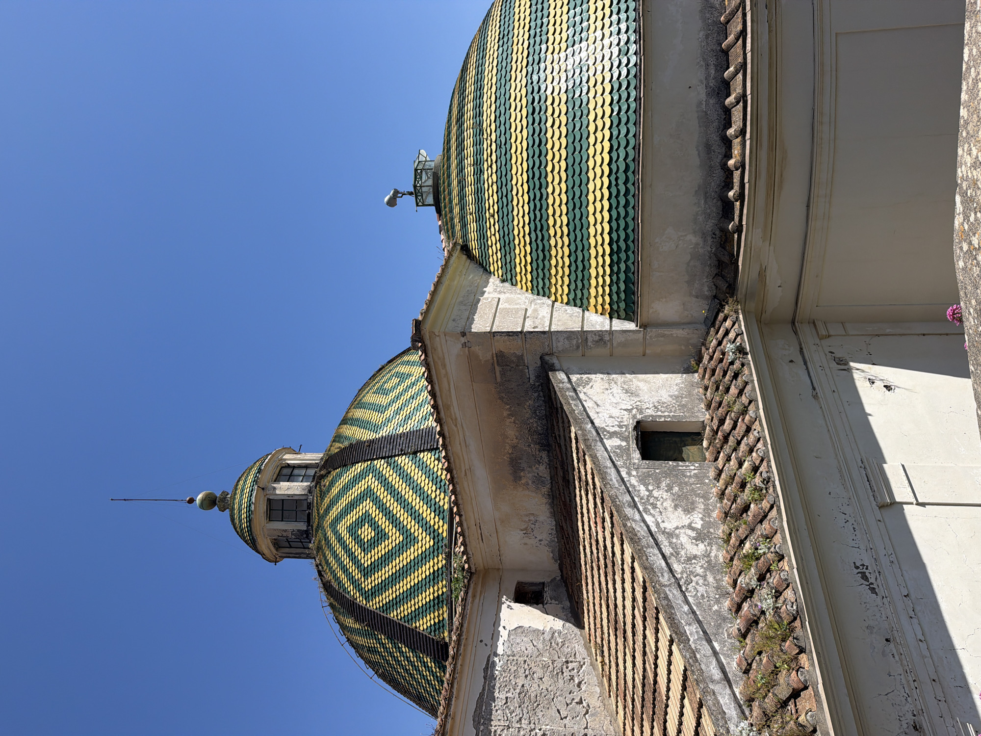 The image depicts the dome of the Collegiata di Santa Maria a Mare church, a landmark in Maiori, Italy. The dome is covered in decorative tiles arranged in geometric patterns. The upper dome features vertical stripes of alternating green and yellow tiles, while the lower dome has a diamond pattern with the same colors. The church architecture is visible, showing the aging facade with textured details, a small window, and some vegetation growing on the surfaces. The background is a clear blue sky, typical of the coastal climate.