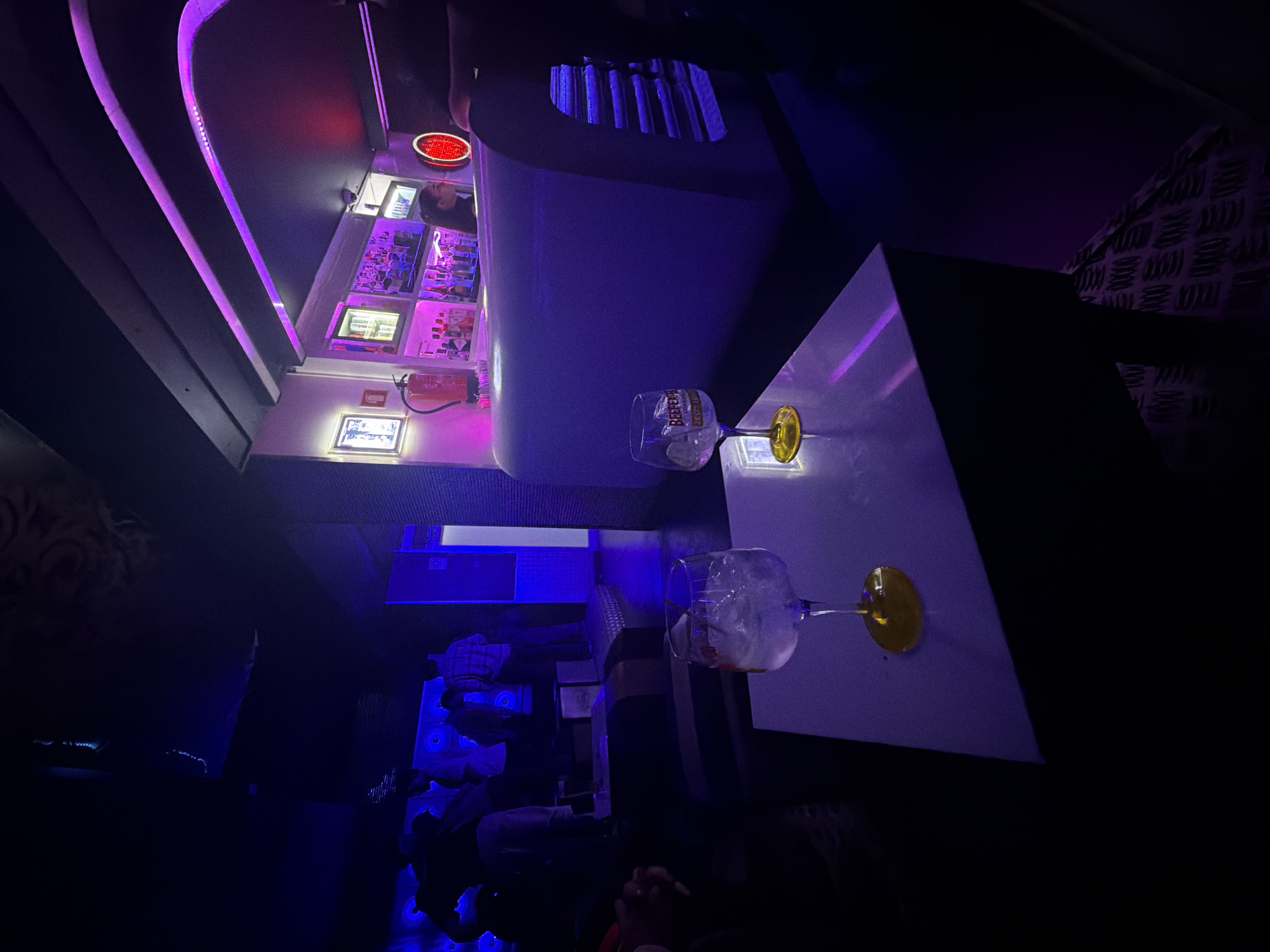 The image shows the interior of a bar, possibly in Vila Real, Portugal, given the context. The scene is dimly lit with blue and purple lighting. Two cocktail glasses, one with a "Beefeater" label, are placed on a white table. Behind the table, there is a bar with shelves displaying various bottles and a red fire extinguisher. The bar area is illuminated by neon lights. Further back in the image, there are people sitting at tables, though they are obscured by the darkness. The overall atmosphere suggests a nightclub or lounge.