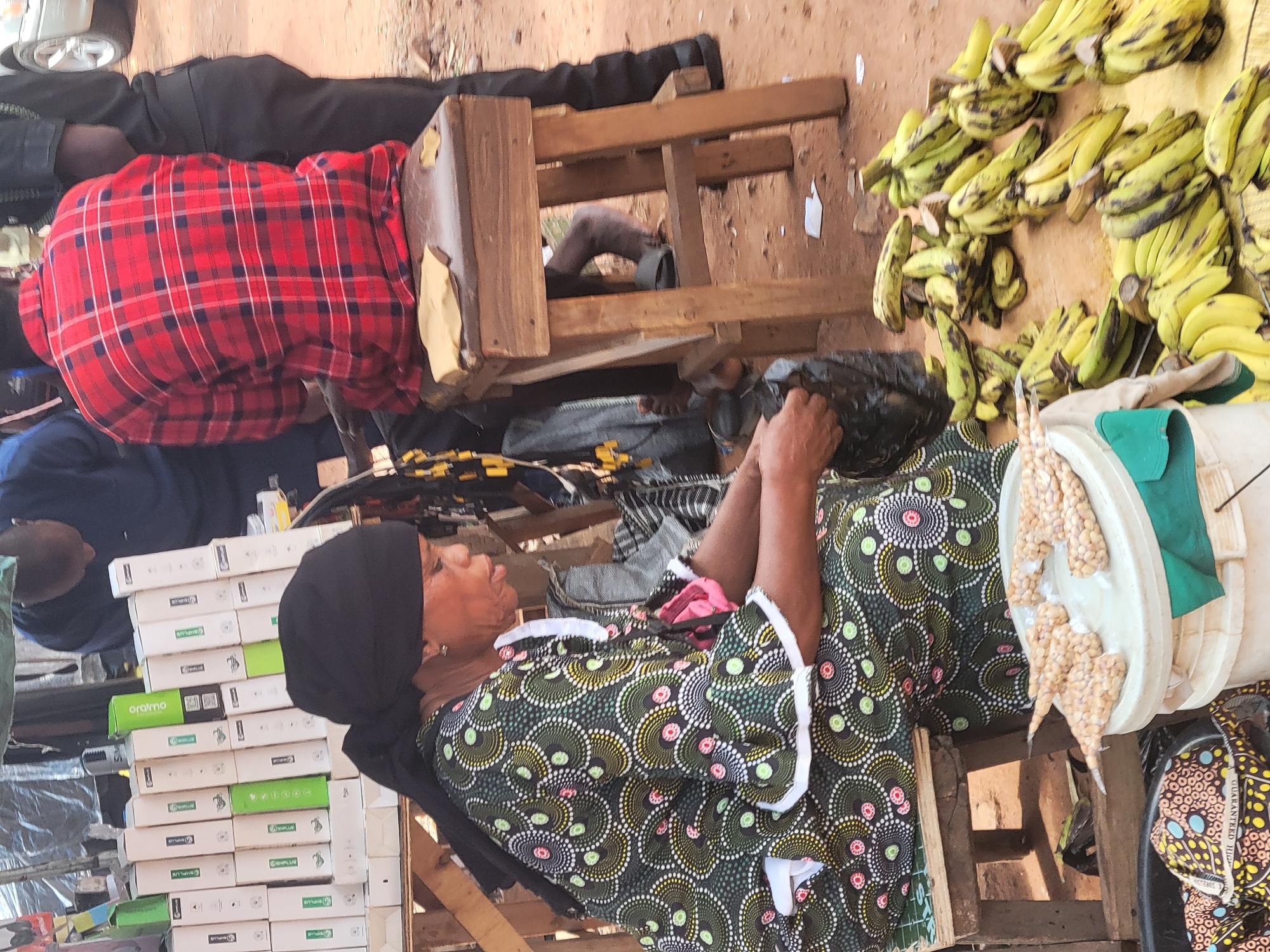 In an outdoor setting in Abuja, Nigeria, an African woman wearing a black head covering and a green and black patterned dress sits on a wooden chair. She is holding a black plastic bag, possibly packing or unpacking items. Next to her is a white container with what appears to be packaged food. In front of her are stacks of white boxes with green accents. To her right, there are bunches of ripening bananas. Nearby, a person wearing a red plaid shirt sits on another wooden chair. The setting appears to be a market. Other people are present in the background.