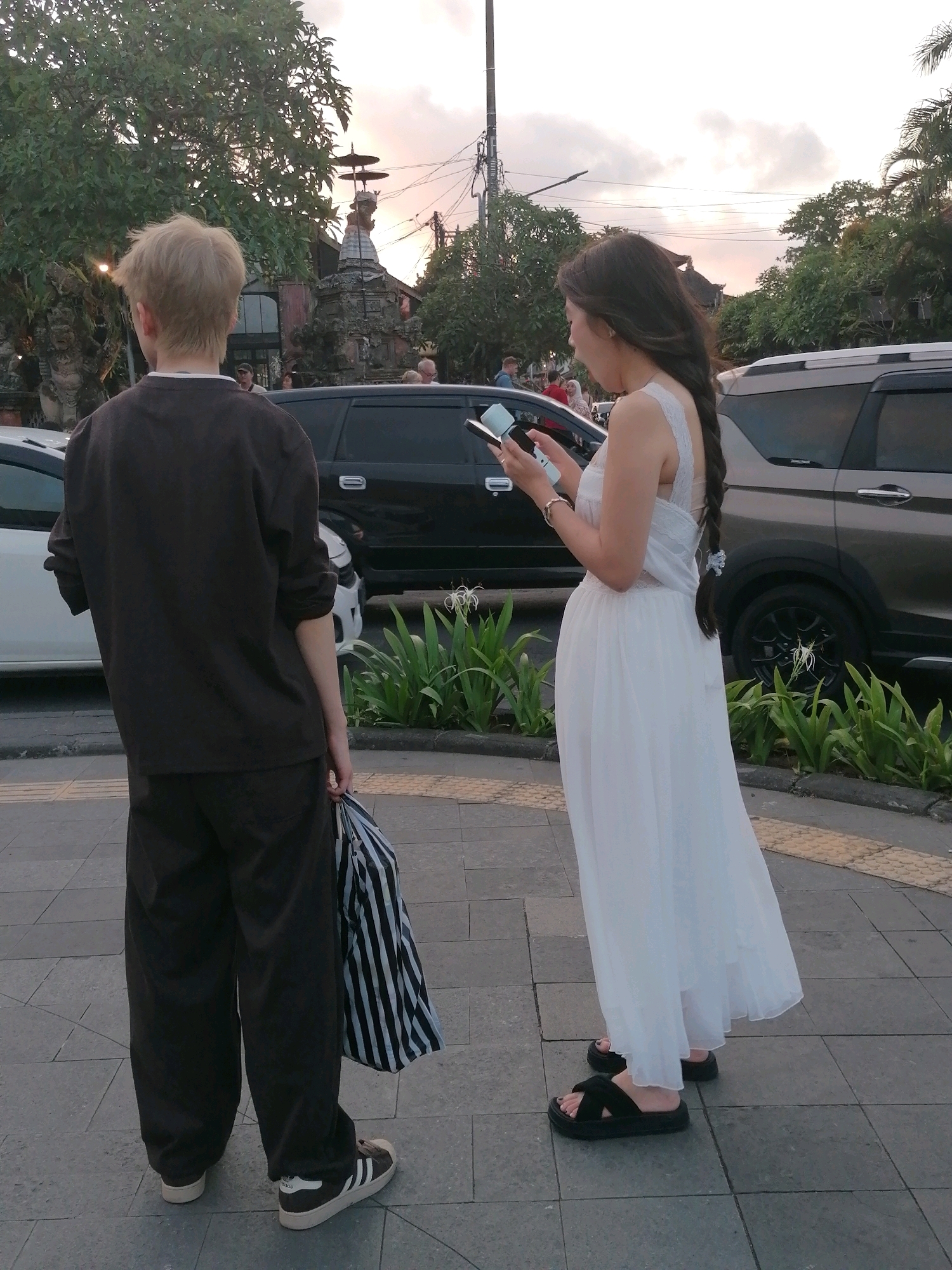 The image shows two people standing on a paved sidewalk in the city of Ubud District, Indonesia. On the left, a person with short, light-colored hair wears a dark brown long-sleeved shirt and loose-fitting dark pants. They are holding a striped bag with black and white vertical stripes in their right hand. On the right, a person with long, dark braided hair wears a white sleeveless dress. They are holding a black smartphone in their left hand and a light blue object, possibly another phone or a portable charger, in their right hand. Both individuals are facing away from the camera, looking towards the street.
Behind them, several vehicles are parked along the road, including a black SUV and a silver SUV. The background features lush green trees, power lines, and a building with traditional Indonesian architecture, including a tiered roof structure. The sky appears overcast with hints of sunset colors. The sidewalk is made of grey paving stones with yellow edging. The scene is outdoors, suggesting a public street or walkway.