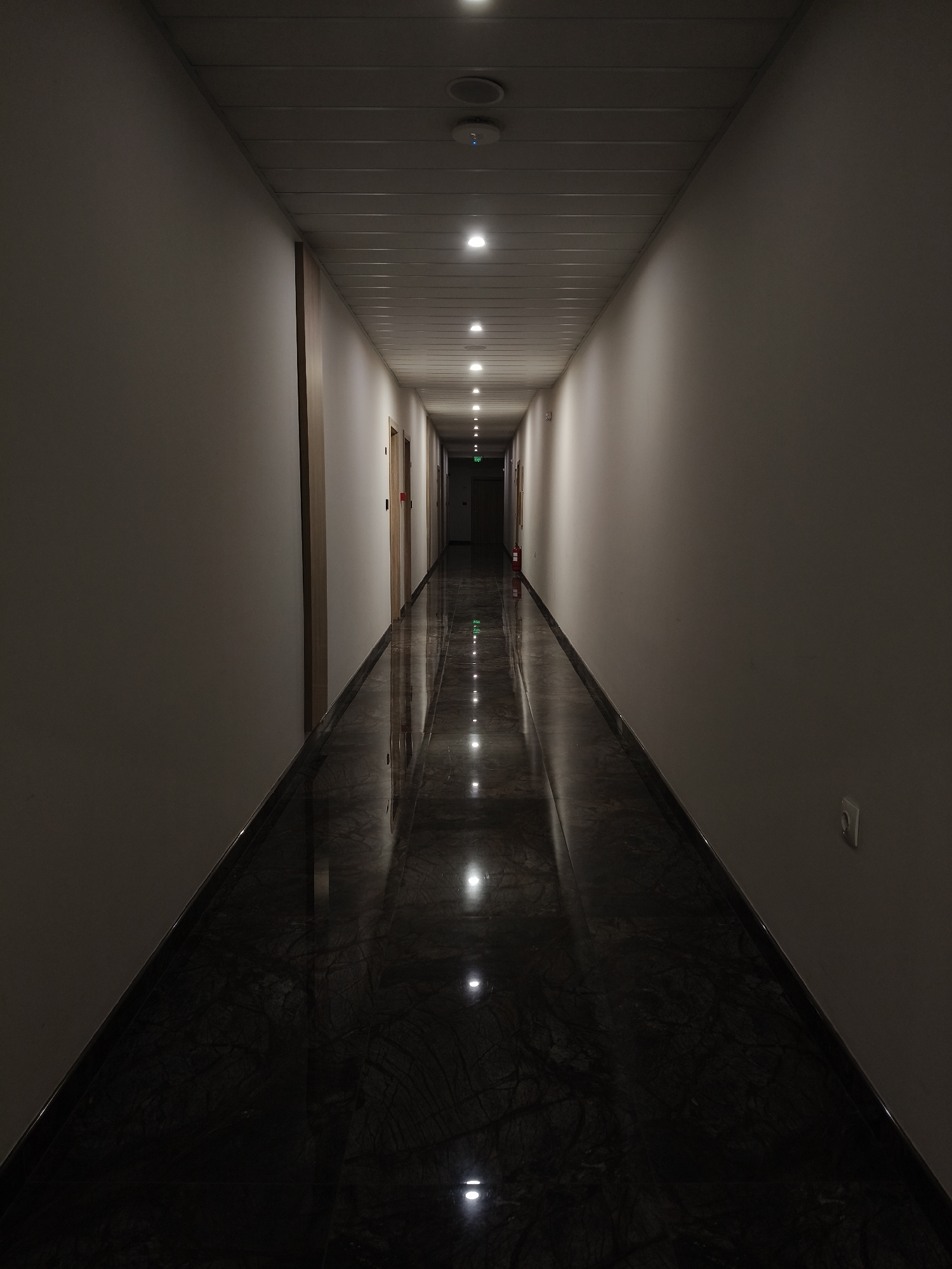 A long, dimly lit hallway with a shiny, dark marble floor. Recessed ceiling lights stretch down the corridor, reflecting on the floor. Doors line both sides of the hallway. The walls are light-colored, and there is an exit sign visible at the far end of the hallway. An electrical outlet is visible on the right wall. There is no indication of the location being in Plovdiv, Bulgaria.