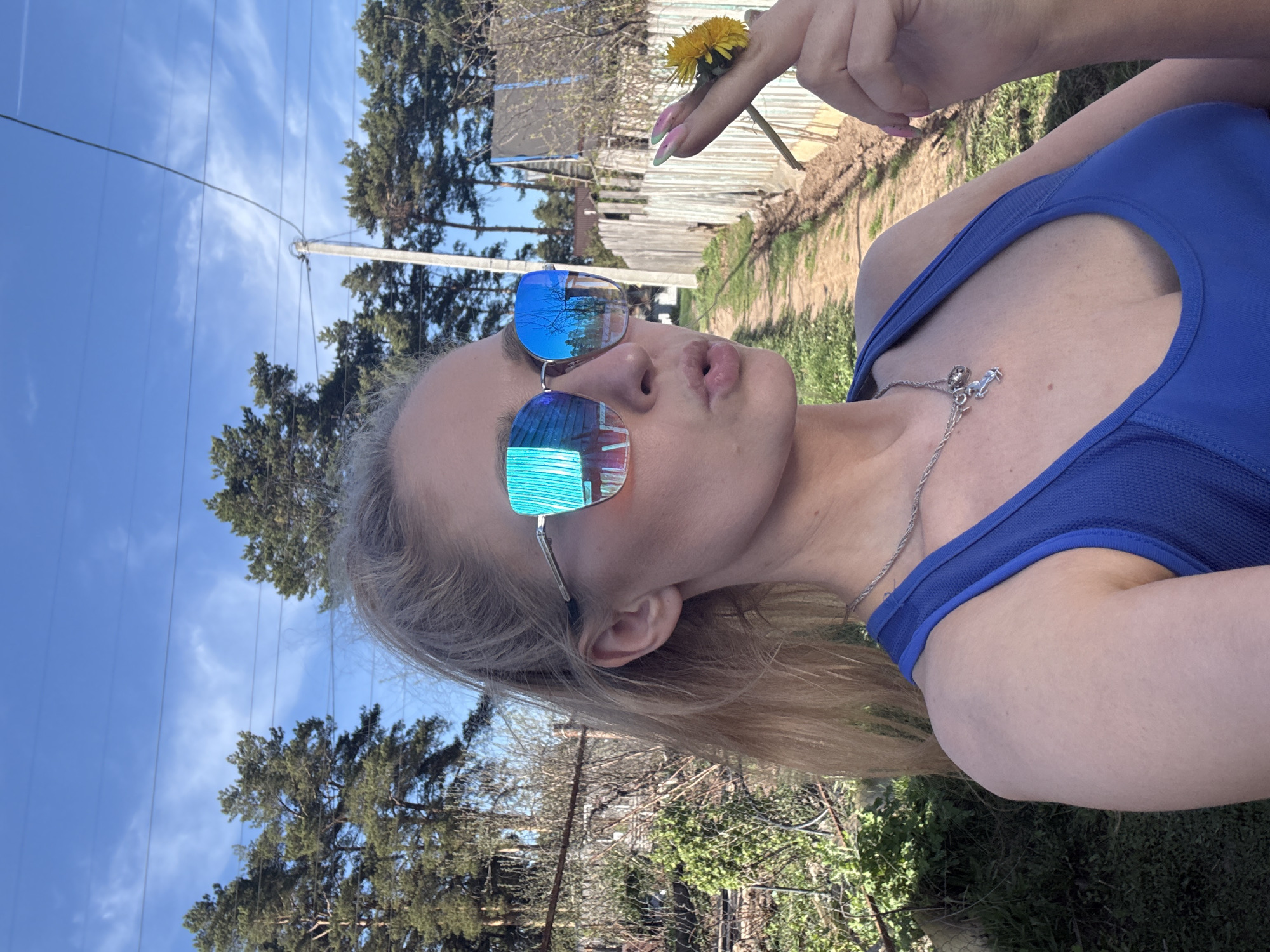 A fair-skinned young woman in a blue tank top is holding a dandelion in her manicured hand. She is wearing reflective sunglasses and a silver necklace. Her hair is blonde. The background shows trees, a blue sky, and a glimpse of a rustic wooden structure. Utility poles and lines are visible. The location is likely a suburban or rural area in Chaikovsky, Russia.