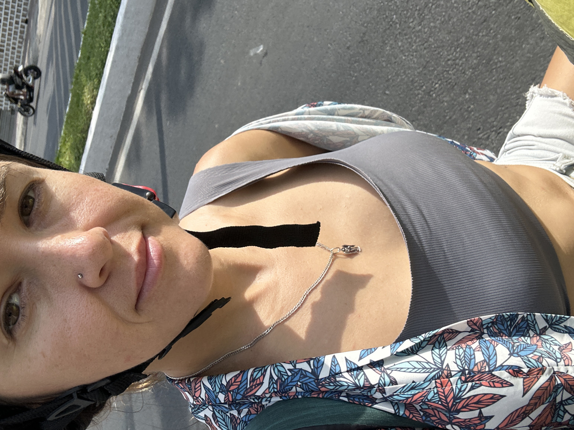 A woman is captured from the chest up, wearing a grey tank top, a patterned open-front shirt with blue, red, and white leaf designs, and a black helmet. A silver necklace with a small angel pendant rests on her décolletage. Her nose is adorned with a small silver stud. The background suggests an outdoor setting, possibly a street in Nha Trang Ward, Vietnam, with a paved surface, a concrete curb, and a patch of green grass. In the distance, two figures on a motorcycle are visible. The lighting indicates a sunny day.