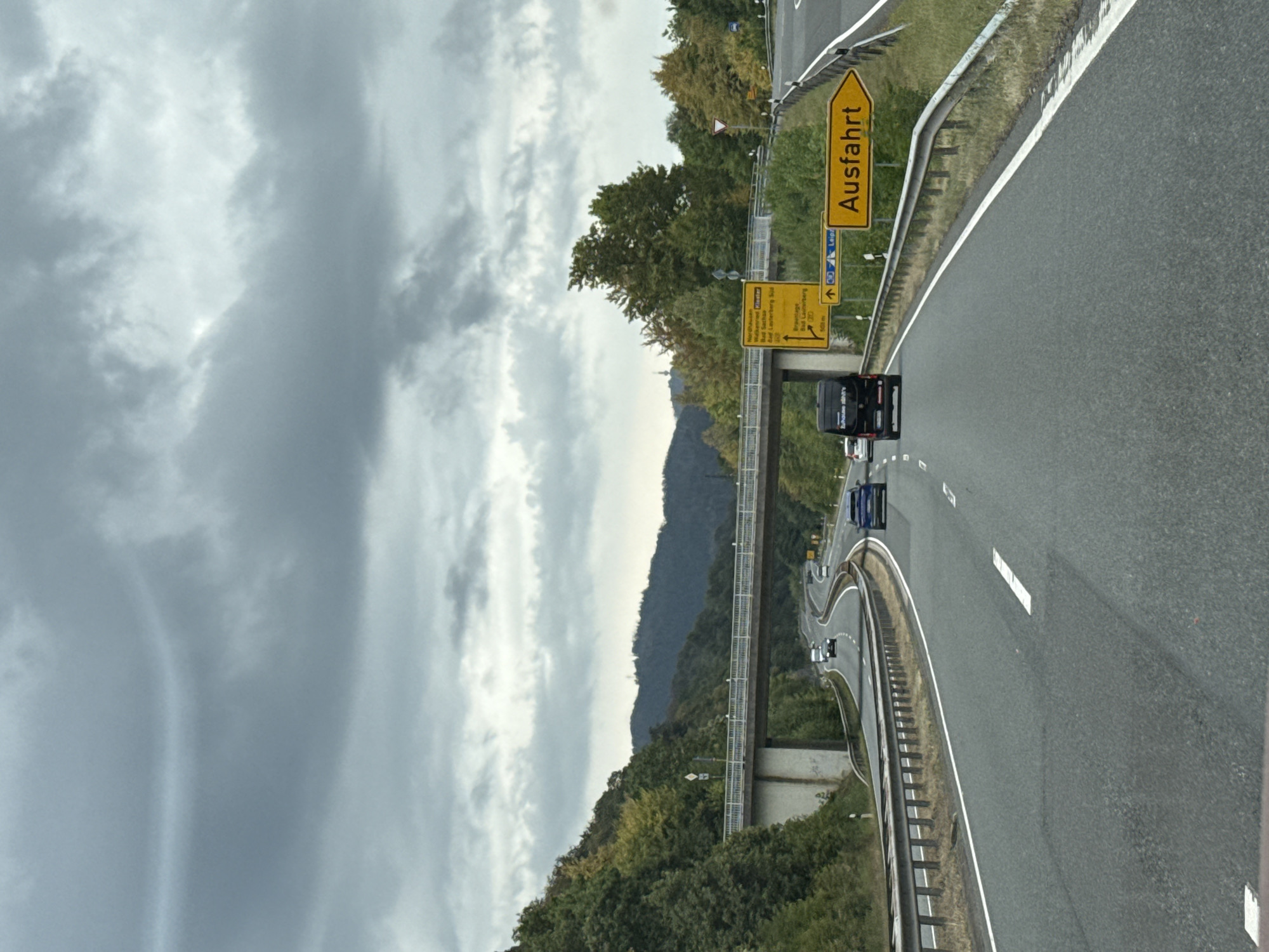 The image shows a view from a vehicle traveling on a highway in Germany, likely in the Harz region of the Göttingen district. The sky is heavily overcast with dark, dramatic clouds. A multi-lane highway curves ahead, with guardrails lining the sides. Several cars are visible on the road, including a dark van and a blue sedan. A pedestrian overpass spans the highway in the distance, supported by concrete pillars. The surrounding landscape is hilly and covered with dense green trees, some showing signs of autumn color. Several yellow road signs are visible on the right side of the highway; one prominently displays the word "Ausfahrt" (Exit) with an arrow pointing to the right. Another sign provides directional information to various towns and cities, including Nordhausen, Bad Sachsa, and Bad Lauterberg, as well as Leipzig. The scene suggests a typical highway drive under cloudy weather conditions in a scenic, rural area of Germany.