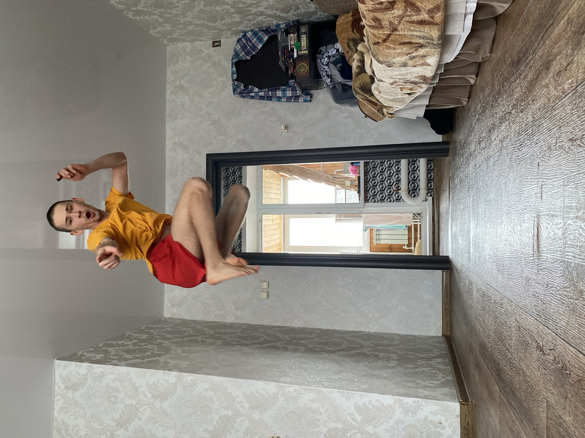 The original image, which is rotated 90 degrees clockwise, captures a dynamic indoor scene in what appears to be a residential setting in Ulyanovsk, Russia. A young man, likely in his late teens or early twenties, is depicted mid-air, suggesting he is jumping or performing an acrobatic move. He has short, light-colored hair and is wearing a yellow short-sleeved t-shirt and red shorts. His mouth is open in an expression of surprise or exertion, and he holds a small, dark object (possibly a marker or pen) in his right hand, pointing with his left. His legs are bent, indicating movement.
The room features light-colored walls adorned with a subtle, patterned wallpaper. The floor, visible on the right side, is covered with wooden laminate flooring. To the upper right, a piece of furniture, possibly a dresser or shelf, is seen, topped with a blue and white plaid shirt, books (one with visible Russian text "ПОДГ" on its spine), and other miscellaneous items. Part of a textured, patterned blanket or throw covering another piece of furniture (like a couch or bed) is also visible.
A dark-framed opening, appearing to be a doorway or large window, leads to an outside area. Through this opening, a white-framed window is visible, along with a wooden building exterior and a white laundry drying rack with clothes hanging on it. The bright light streaming in suggests it is daytime. Two white electrical outlets or switches are mounted on the wall near the bottom of the dark frame.
