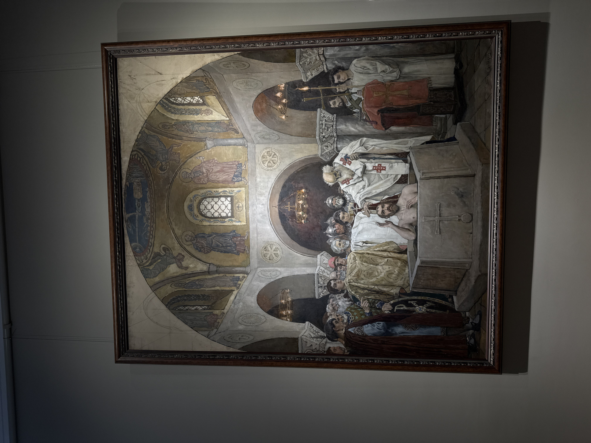 The image depicts a large, framed painting hanging on a wall, likely in a gallery or museum. The painting shows a scene of a religious ceremony, possibly a burial or anointing, taking place within a church or cathedral setting. The background is filled with Byzantine-style frescoes and architectural details, including arches, windows, and decorative elements. A group of individuals, including priests in ceremonial robes and other figures, surround a partially exposed figure lying on a stone slab. The overall palette of the painting consists of earthy tones and muted colors. The scene has a solemn and reverent atmosphere. The painting is titled "The Lamentation of Christ" by Boris Kustodiev, and it can be found in the Nizhny Novgorod State Art Museum, Russia.