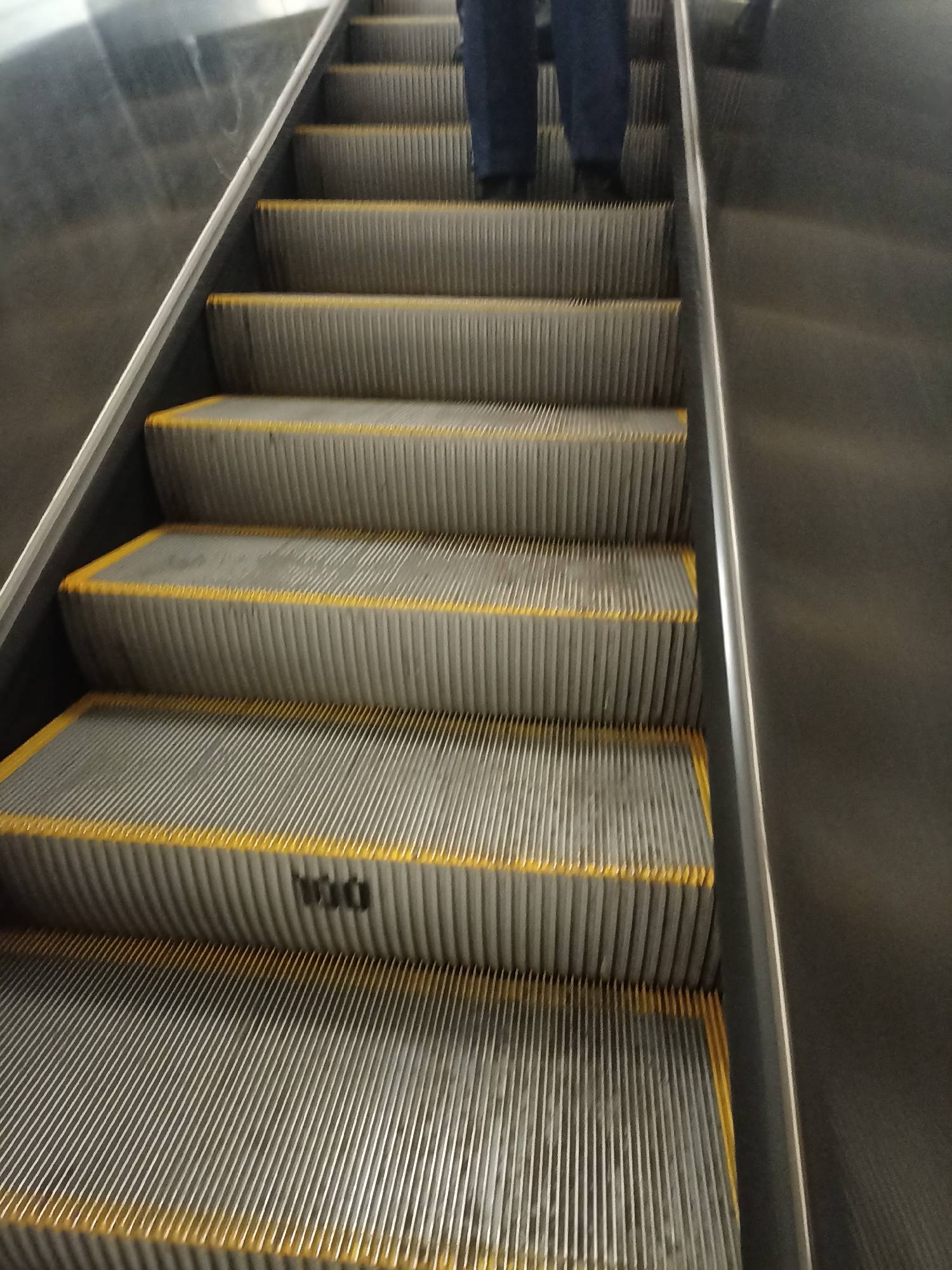 The image shows an upward-facing perspective of an escalator. Several steps of the escalator are visible, each with textured metal treads and bright yellow safety strips along the edges. A person's legs, clad in dark blue trousers and black shoes, are positioned on the upper steps, indicating they are riding the escalator upwards. To the right of the escalator steps is a dark, reflective metallic surface, likely part of the escalator's housing or a surrounding wall. The overall environment appears to be an indoor public space, possibly a metro station or shopping mall. The image does not provide specific clues to pinpoint a location within Moscow, Russia.