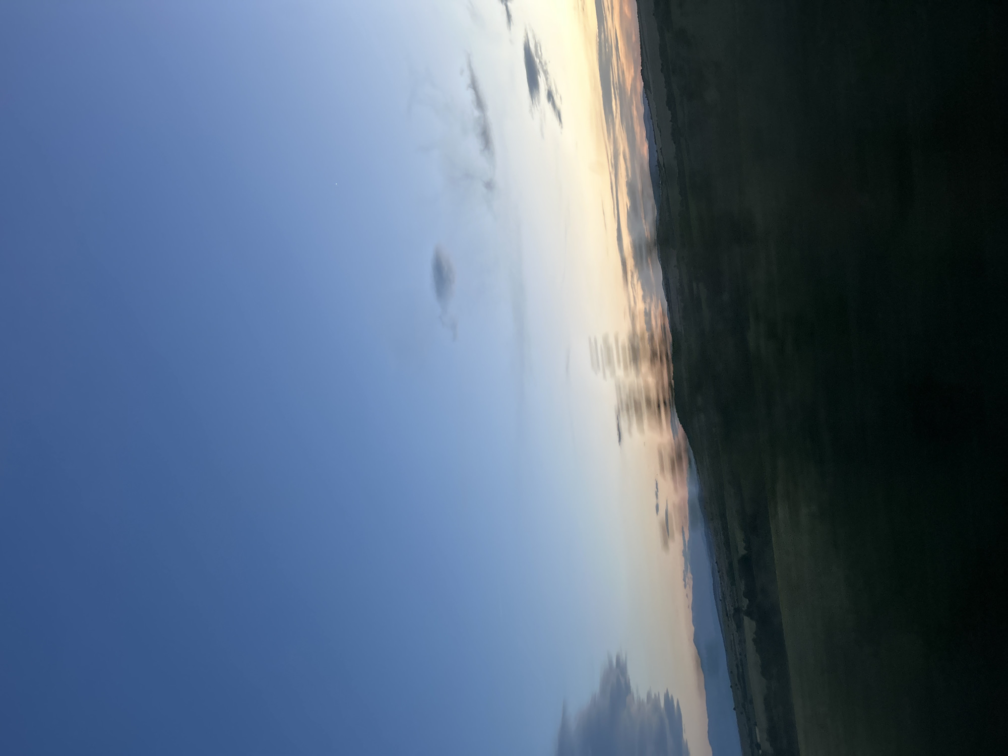 A landscape scene captured in Smilets, Bulgaria, reveals a sky transitioning through sunset. The upper left is a clear blue, slowly merging into bands of yellow, orange, and grey-toned clouds as the sun approaches the horizon. Below this, a blurred line indicates the top of a dark mass, possibly representing land or water reflecting the diminishing light. The ground vegetation or landscape is nearly impossible to ascertain. There are no people visible in the image. The location is Smilets, Bulgaria.