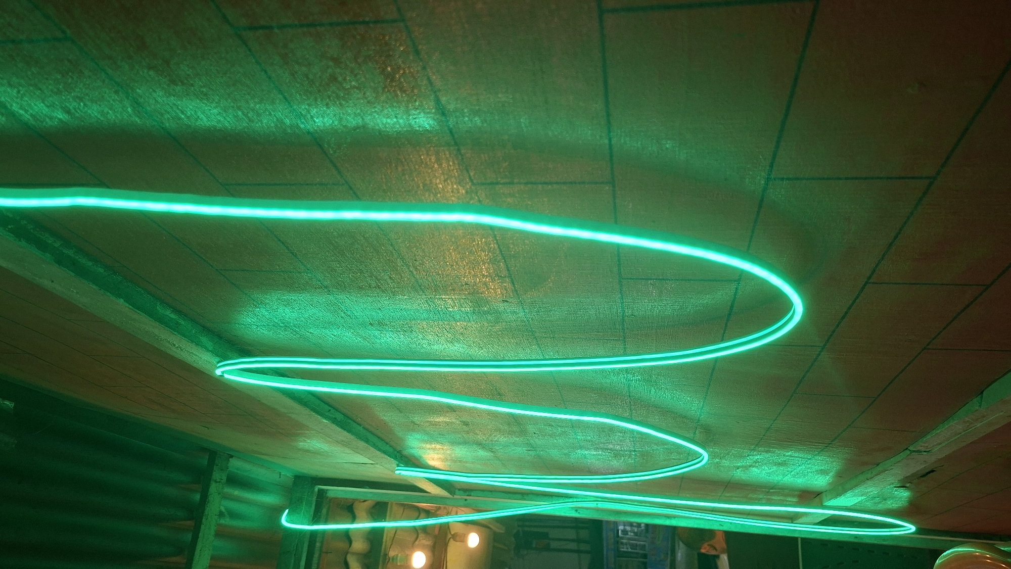 A neon green light tube forms abstract wavy lines on a brick wall. The light is bright and casts shadows on the wall, giving it a textured appearance. The wall is made of reddish-brown bricks with dark grout lines. Above the wall, a corrugated metal roof is visible. In the background, on the left side of the image, there are lights and a person visible, and some equipment that looks like it might be part of a construction site. The image appears to be taken indoors, possibly in a bar or restaurant, given the neon lights. The overall impression is one of modern, artistic decoration with a vibrant, energetic feel.