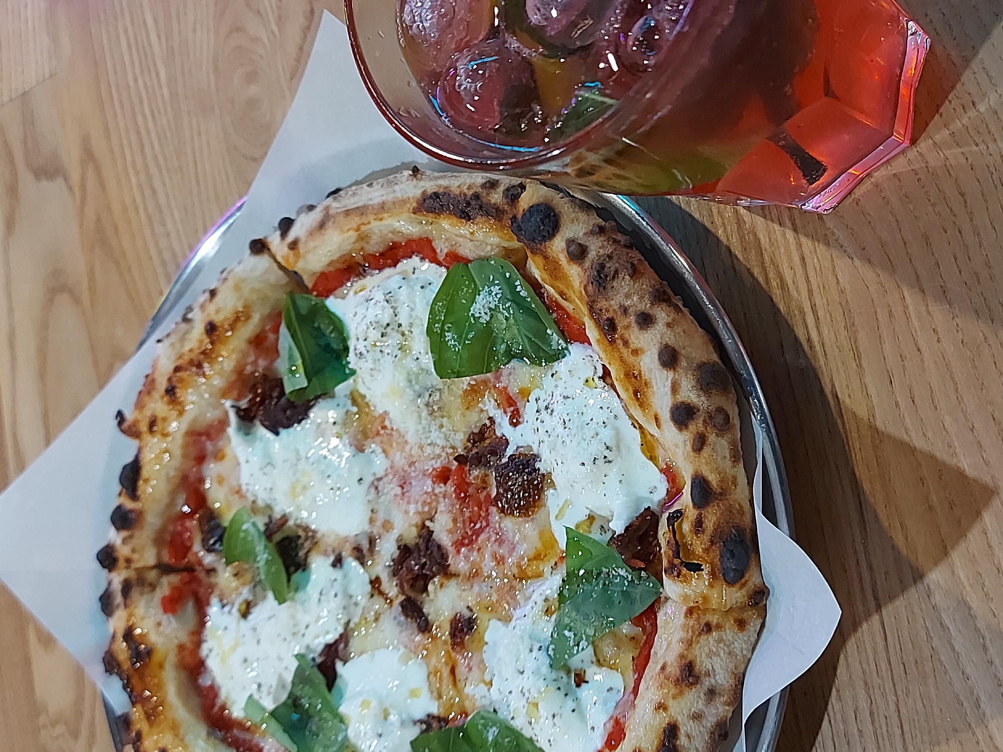 A high-angle close-up shot showcases a pizza served on a metal tray with white paper, next to a glass filled with a red beverage and ice. The pizza features a charred crust, topped with red sauce, chunks of what appears to be sun-dried tomatoes, dollops of fresh mozzarella, and basil leaves. The drink in the glass is a bright red color with ice cubes and garnish, possibly lime or mint. The background is a light wooden surface. Location details about Saint Petersburg, Russia are not discernible from this image.