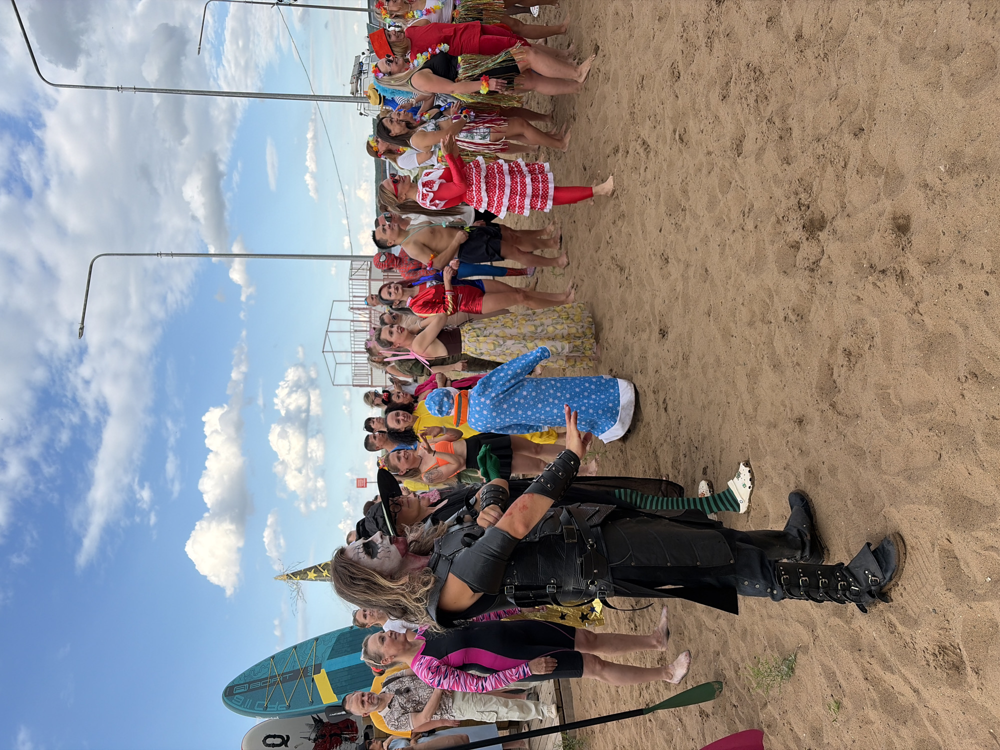 A group of people in costumes are gathered on a sandy beach, possibly for an event. The scene appears to be taking place on a sunny day with a blue sky and scattered clouds. In the background, several streetlights are visible, and further back, a section of a white structure with railings can be seen.
Several individuals are dressed in various outfits. One person in the foreground is wearing a dark, elaborate costume resembling armor, complete with buckles and straps, and their face is painted with white and dark makeup. To their right, another person wears a black witch's hat. Further into the crowd, people are dressed in colorful clothing, including what appears to be a Spider-Man costume, Hawaiian-themed attire with leis and grass skirts, and red and white polka-dot outfits. There are also several surfboards standing upright in the background.
The ground is sandy, with footprints visible, indicating recent activity. The overall impression is of a lively outdoor gathering or festival. The location is specified as the beach in Chaikovsky, Russia.