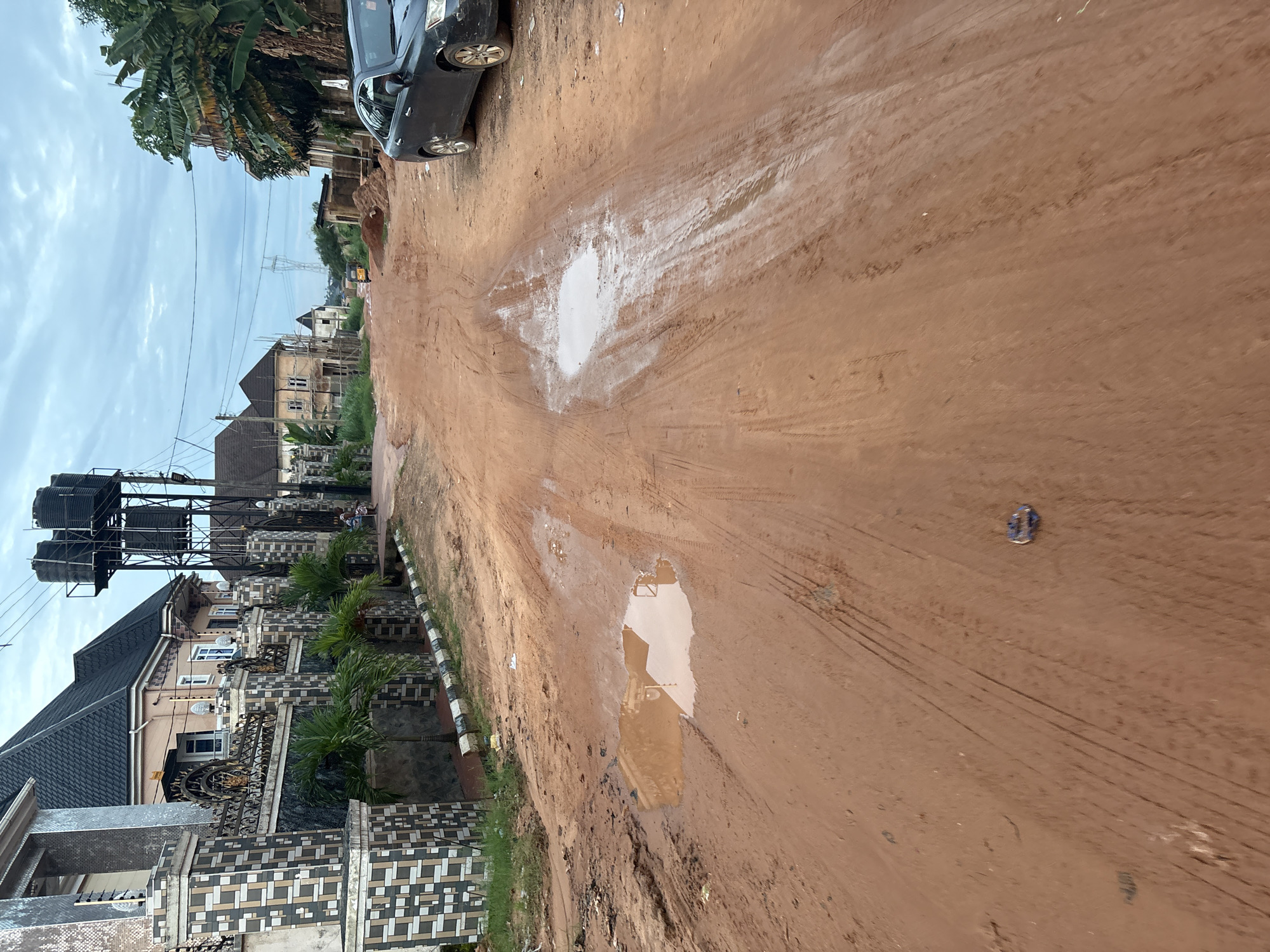 This image shows a dirt road in Obosi, Nigeria, with a car on the right side and houses on the left. The road is muddy with tire tracks visible, and there are puddles of water on the ground. A water tower is visible in the background, along with power lines and some vegetation. The sky is cloudy, suggesting it may have recently rained or is about to. The houses appear to be a mix of modern and under construction, with some featuring decorative fences and tiled roofs. There are a few scattered debris and objects on the road.