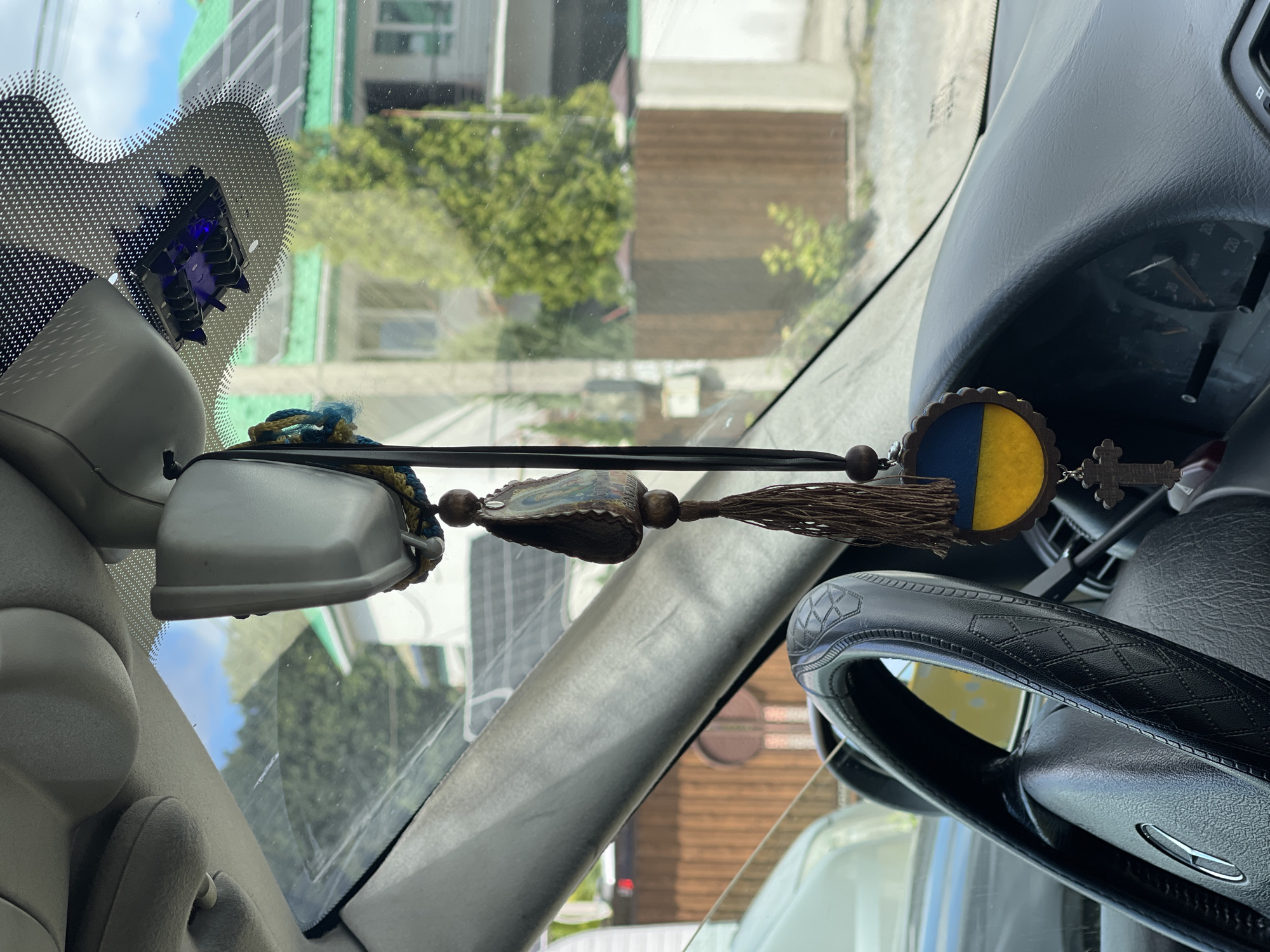 The image is a close-up, interior view of a car in Konopnytsia, Ukraine, focusing on the rearview mirror area and the dashboard. A black steering wheel with a patterned grip is visible on the left. Hanging from the rearview mirror assembly are several car charms. These include a decorative beaded tassel, a small oval pendant depicting an image of a religious figure, and a circular pendant with the blue and yellow colors of the Ukrainian flag. Below the Ukrainian flag pendant, a small wooden Christian cross is also hanging. The car's dashboard and windshield are visible, with a view of houses and trees through the windshield. The lighting suggests it is daytime.
