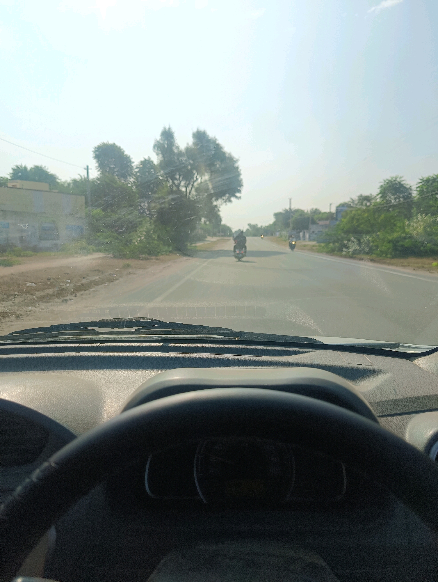 The image is taken from inside a car, looking forward through the windshield. The view is of a paved road with a slight curve to the right. On the left side of the road, there is a dirt shoulder with some scattered debris and vegetation. Further back on the left, there are buildings and trees. On the right side of the road, there is a line of trees and bushes. The sky is bright and hazy, suggesting a clear day. There are at least three motorcycles visible on the road ahead. The dashboard of the car and a portion of the steering wheel are visible in the foreground. The scene appears to be a rural or semi-rural area. The text on the speedometer is illegible. Based on the context provided, this scene is located in Chhapri Bari, India.