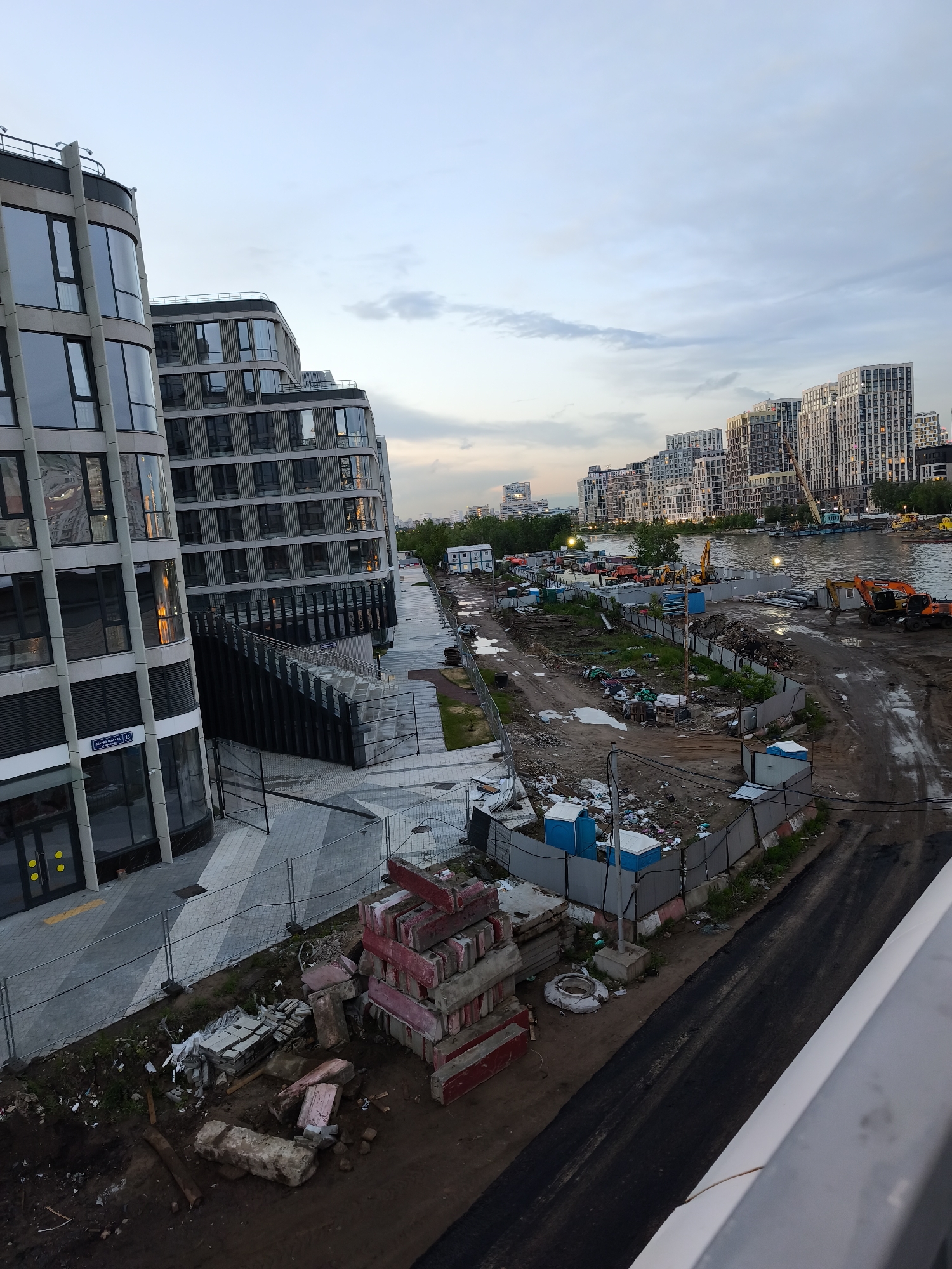 The image shows an urban construction site in Moscow, Russia, bordering the Moskva River. Modern residential buildings with curved facades stand on the left, while a cluster of taller, rectangular apartment buildings can be seen across the river in the background.
The construction area is muddy and cluttered with building materials, including stacks of red and gray concrete blocks, debris, and several portable toilets. Heavy machinery, such as excavators, is present on the riverbank. A fence separates the construction site from a paved walkway adjacent to the new buildings.
The sky is overcast, indicating either early morning or late evening. A street sign "MARIA ULJANOVOY 15" is visible on the building to the left.
