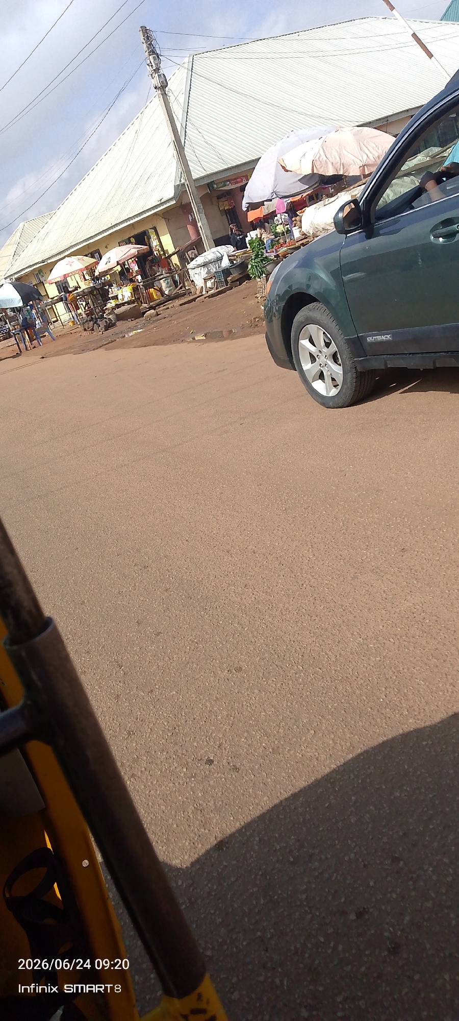 This image captures a street scene in Jos, Nigeria, likely from the perspective of a tricycle. In the background, a row of market stalls with corrugated iron roofs line the street, shaded by numerous umbrellas. Various goods are displayed, and a few individuals can be seen amidst the stalls. A dark green Subaru Outback is parked on the right side of the road, its rear passenger door and wheel visible. The road surface is paved with asphalt. The timestamp "2026/06/24 09:20" and "Infinix SMART 8" are visible in the lower left corner, indicating the date and time the photo was taken, as well as the device used. The overall impression is a bustling local market area on a bright, possibly cloudy day.