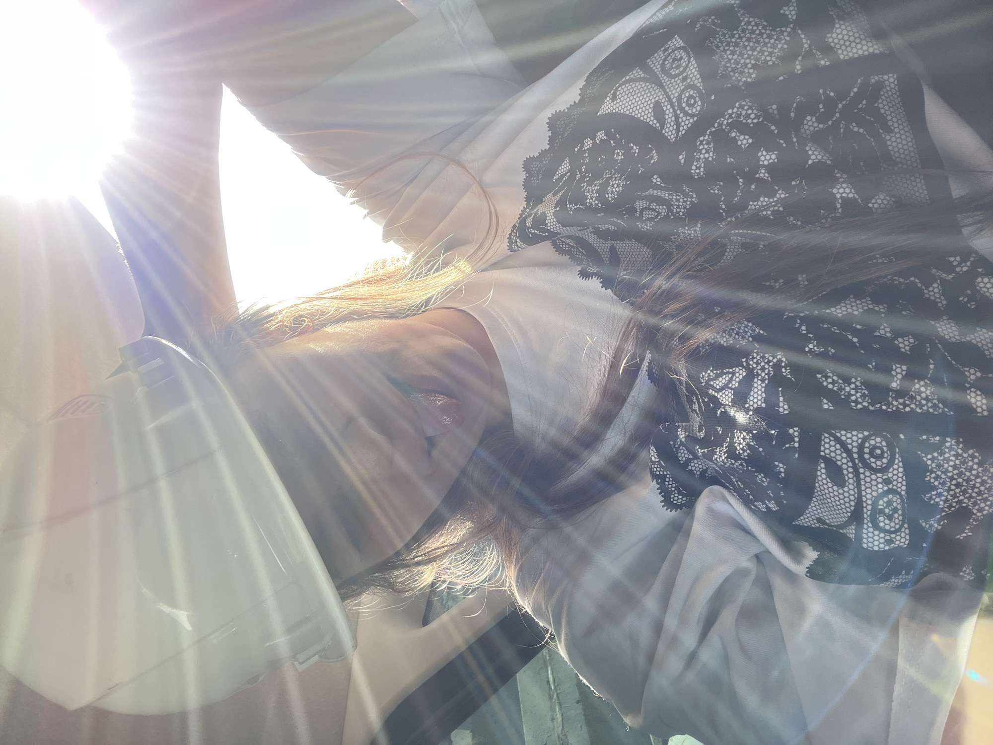 An individual is depicted from a low angle within a vehicle interior, located in the city of Chaikovsky, Russia. The subject wears a white hard hat and a white t-shirt adorned with a prominent black lace-pattern design across the chest. Long, brown hair is visible, illuminated by direct, intense sunlight entering from the upper right, which causes significant lens flare and overexposure, obscuring parts of the face. The individual's tongue is partially extended from the mouth. An arm is raised upwards towards the light source. Indistinct elements consistent with a vehicle interior, including a window frame or pillar, are visible on the left side of the image.