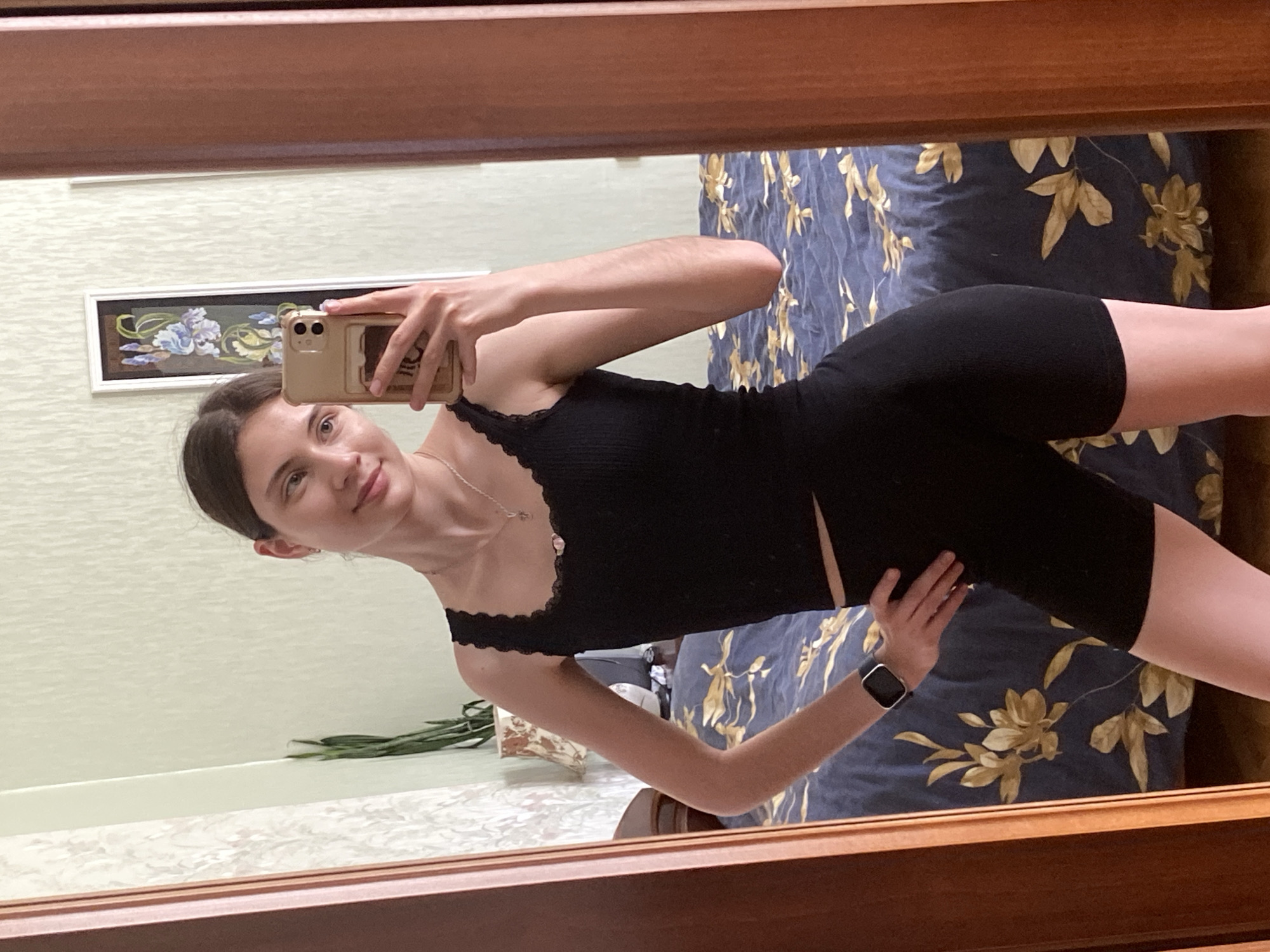 A young woman with brown hair tied back takes a selfie in a mirror. She is wearing a black ribbed tank top with lace trim and black biker shorts. She has a silver necklace with a small pendant and a black smartwatch on her left wrist. The background appears to be a bedroom with a patterned blue and gold bedspread and a framed artwork on the wall. The mirror is framed by a wooden border.
