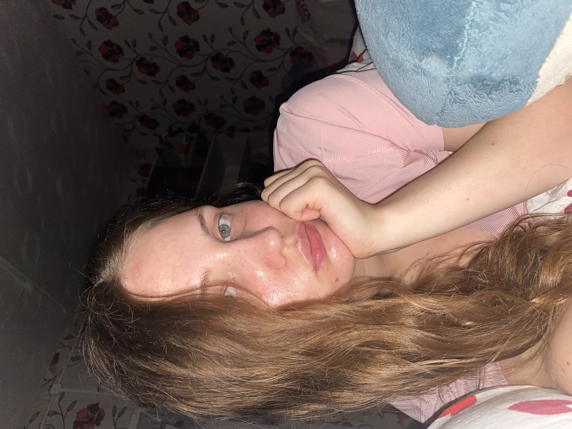 The image shows a close-up of a young woman's face, likely taken indoors at night. She has long, wavy brown hair and is wearing a pink shirt. Her left hand is propped under her chin, and her right arm is visible. The background features floral wallpaper and a dark, indistinct area that could be furniture or a wall. The lighting is dim, and the woman's skin appears slightly oily. There is no indication of the city of Вассята, Russia, or any urban environment. The scene is intimate and personal, suggesting a casual or private setting.