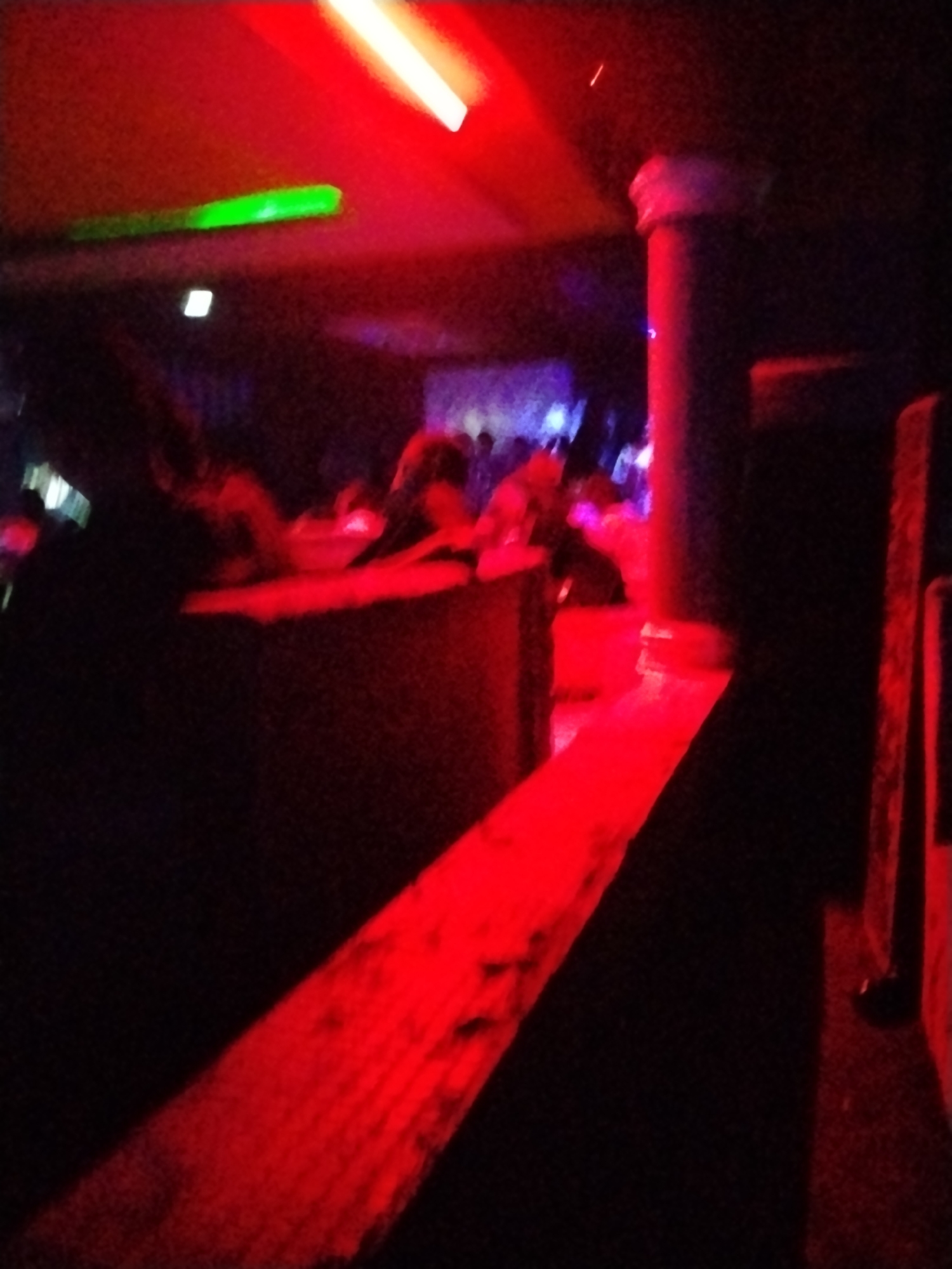 The image depicts an interior scene illuminated by red and green lights. In the foreground, a red-lit surface with a textured pattern extends diagonally from the bottom left corner towards the center. To the right, a dark, cylindrical object, possibly a pillar, is visible, also reflecting red light. In the middle ground, there appears to be a raised platform or counter with a dark, rough surface. Behind this, a group of people are faintly visible, some in silhouette or partially obscured by the dim lighting and red hues. The overall impression is of a dimly lit indoor space, potentially a nightclub or entertainment venue, with vibrant colored lighting creating a dynamic atmosphere. The scene is undated and no specific location within Durumi, Nigeria can be identified from the visual information provided.
