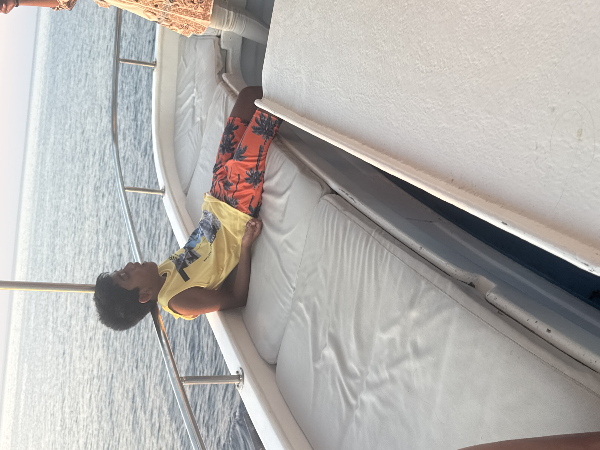 The image shows a young boy reclining on a white cushioned bench on the deck of a boat. He is wearing a yellow tank top and orange shorts with a palm tree pattern. The boat is on the water, with a railing in the foreground and a metallic pole supporting a canopy, partially visible. The sky is a soft pinkish hue, indicating either sunrise or sunset. The water is calm and a muted blue. The boat appears to be in the sea. The location is the city of Polis Chrysochous, Cyprus, as the image was captured in this vicinity. There is another person's legs visible at the far right of the image, wearing light-colored trousers.