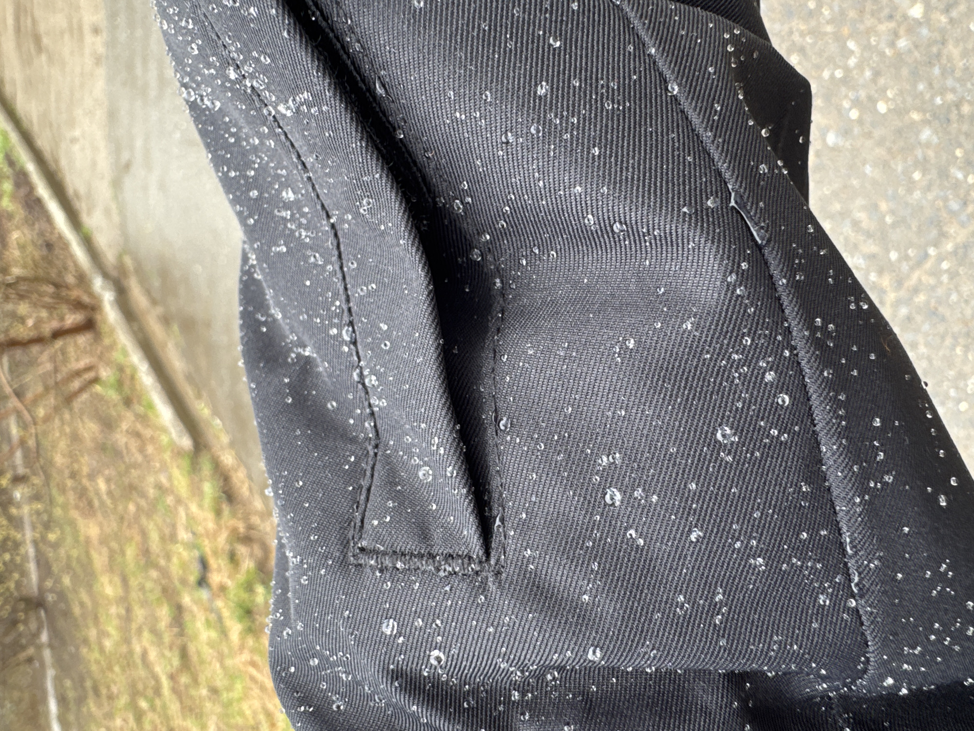 The image shows a close-up of a dark gray or black textured fabric, possibly clothing, covered in water droplets. The fabric appears to be of a twill or ribbed construction. There's a visible seam or pocket flap detail. The background is blurry but suggests an outdoor setting with a concrete or paved surface and some vegetation. There is no evidence to suggest the location is in the city of Chaikovsky, Russia.