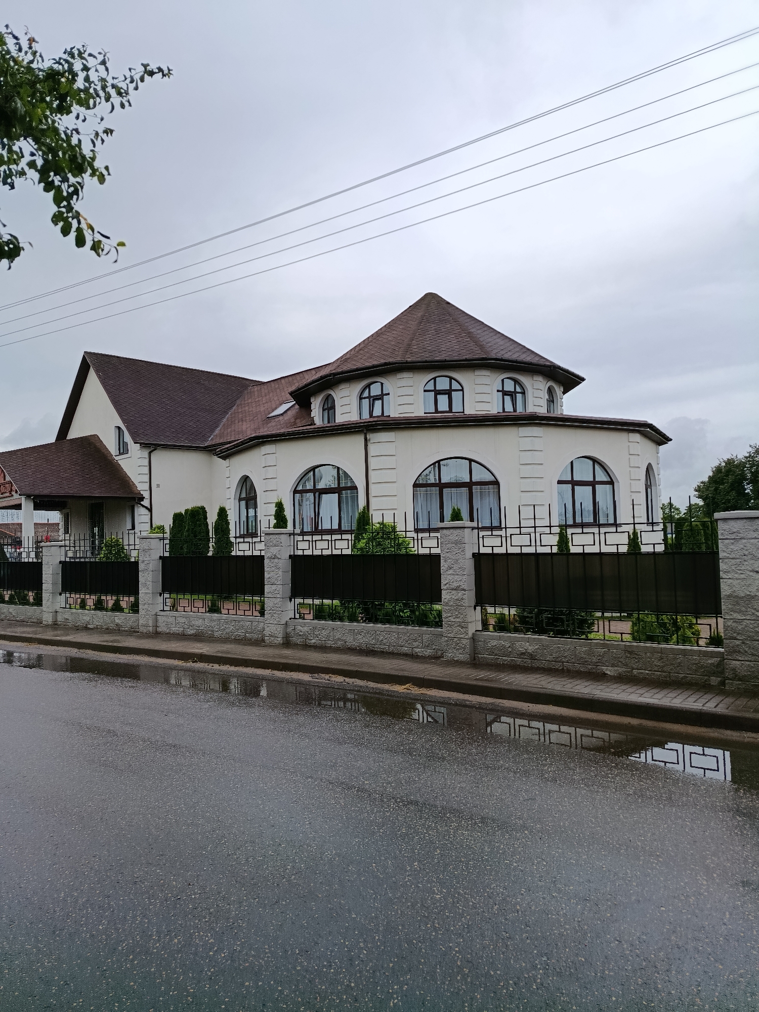 The image shows a modern, multi-story house with a white facade and a brown tiled roof in Смілавіцкі сельскі Савет, Belarus. The house features a circular section with arched windows on the upper floor and larger arched windows on the ground floor. A black metal fence with decorative stone posts separates the property from the street. The street in front of the house is wet and reflective, indicating recent rain, with puddles visible. Overhead, power lines stretch across the overcast sky, partially obscured by the branches of a tree on the left side of the frame. The overall environment appears to be a residential area on a cloudy day. There are no people visible in the image.