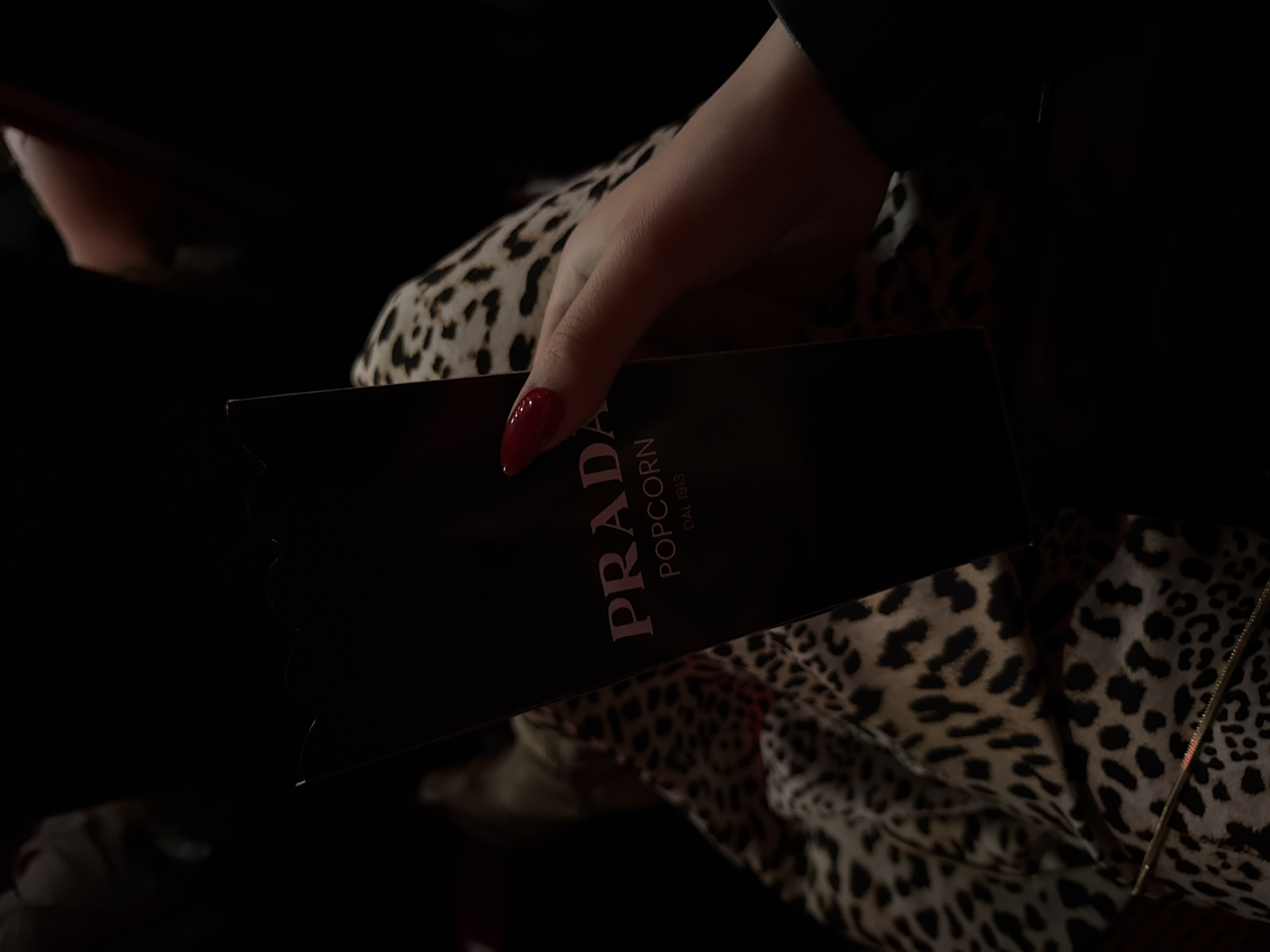 The image shows a hand holding a black container of "PRADA POPCORN DAL 1913". The background appears to be a leopard print fabric. The setting is dark, possibly a movie theater. There is no information that would help locate this image in Yerevan, Armenia.