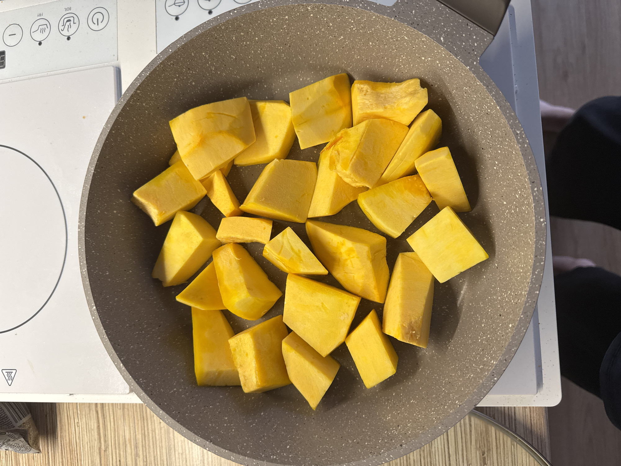 The image shows a close-up overhead view of a grey speckled non-stick frying pan filled with cubed bright yellow pumpkin pieces. The pan is placed on a white electric induction hob. To the left of the pan, a wooden countertop is visible. In the background, a corner of a bag with text and a symbol indicating heat is visible. The scene appears to be a domestic kitchen. There is no discernible location in the city of Perm, Russia, as the image lacks any identifying landmarks or contextual clues. The image focuses on the food preparation process.