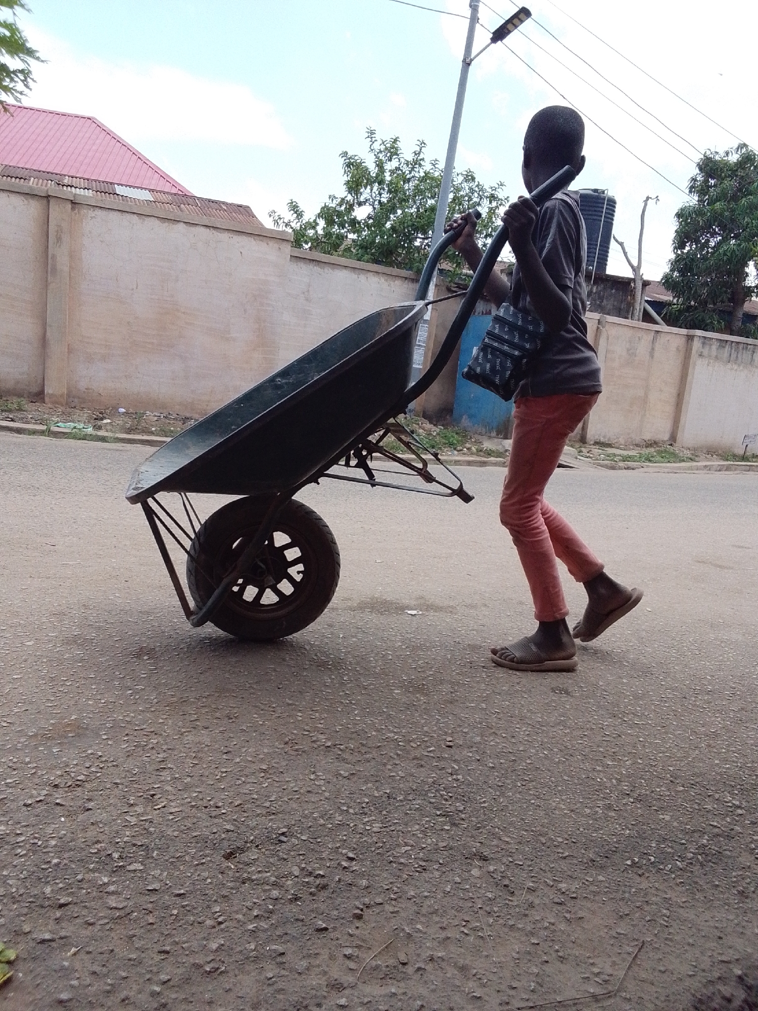 A young person is seen pushing a wheelbarrow on a paved road. The individual is wearing a dark shirt, red pants, and sandals. They have a bag slung over their shoulder. The wheelbarrow is tilted slightly upward.
In the background, there is a wall and a building with a red roof. A utility pole is also visible. Trees are also present in the background. The location is in the city of Unknown, Unknown.
