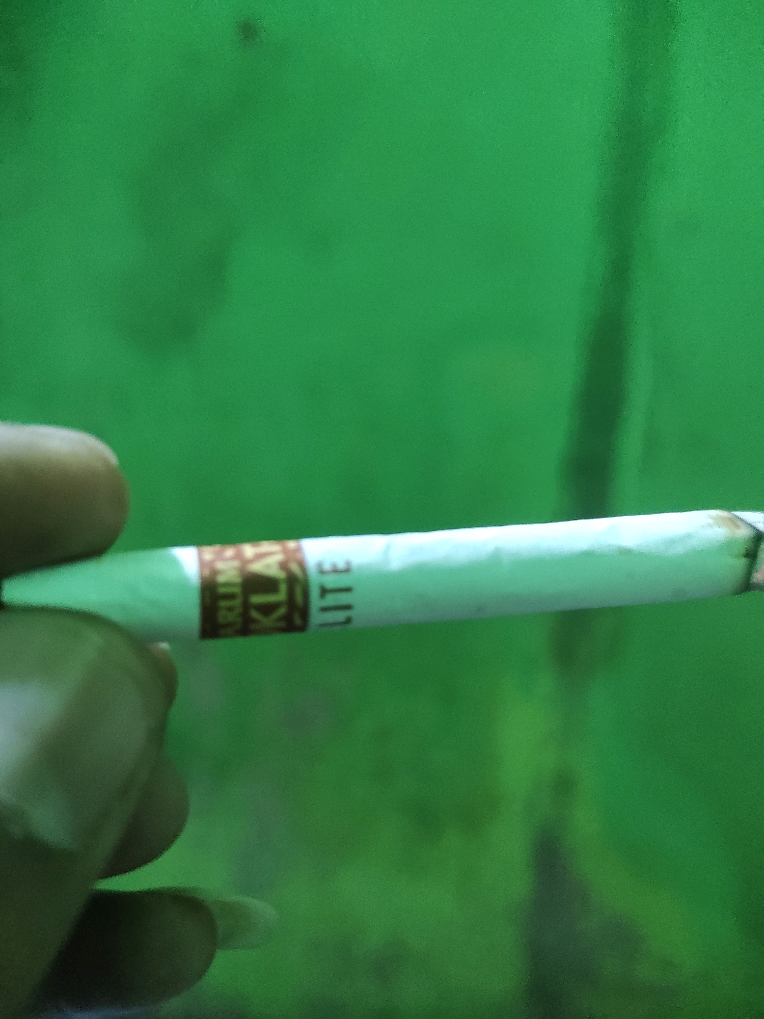 A person's hand is holding a cigarette. The cigarette has a white filter and a brown band with text on it. The background is a blurry green color, possibly a wall or fabric. The image appears to be taken indoors or in a shaded area. The person's fingers and fingernails are visible, suggesting a close-up shot. The overall lighting is dim, contributing to the blurred effect. The location is identified as Padalarang, Indonesia.