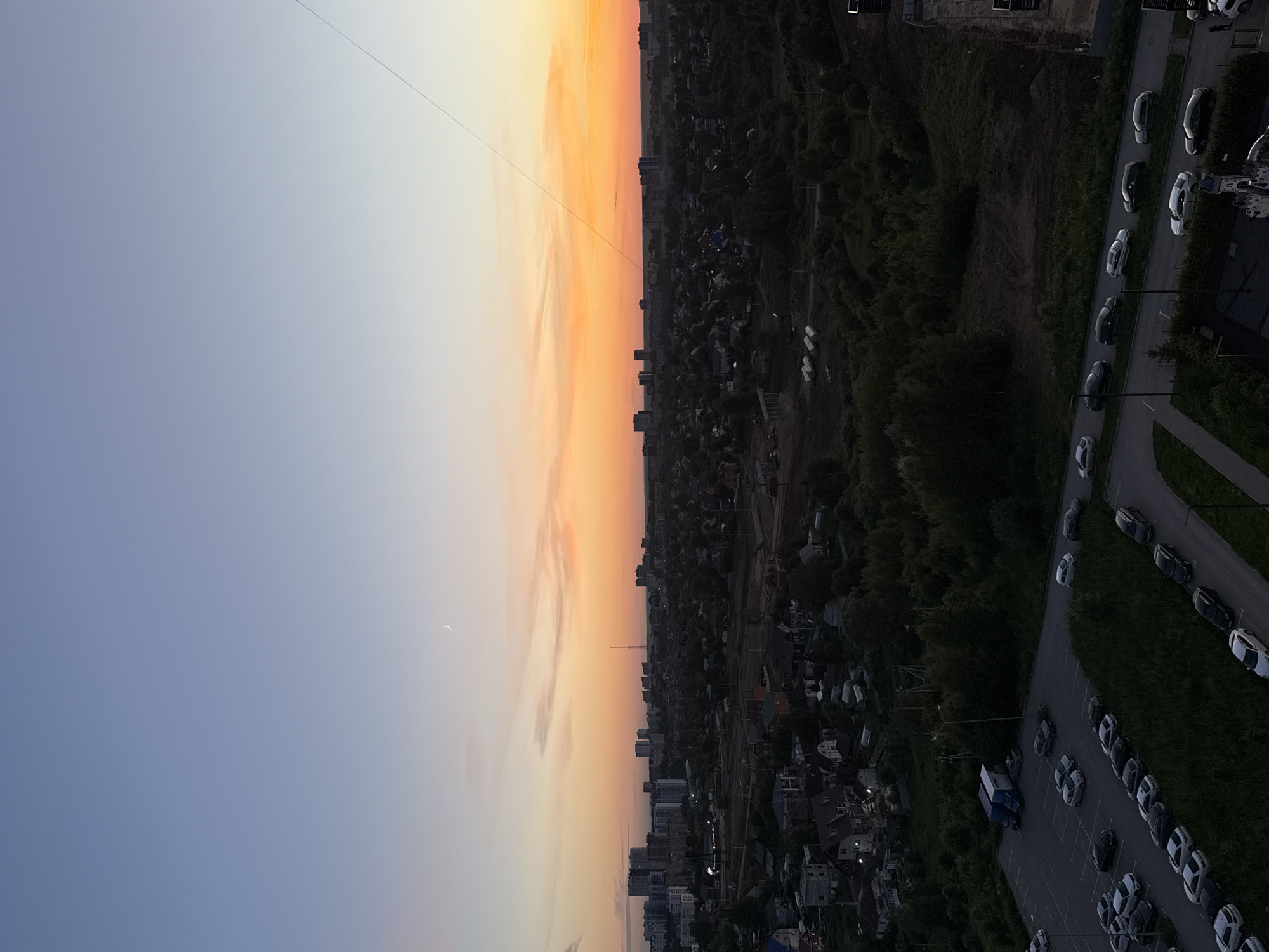 The image depicts an aerial view of a city at dusk. The sky transitions from a pale blue at the top to a vibrant orange and yellow near the horizon, indicating sunset. A crescent moon is visible in the upper left portion of the sky. The city below consists of a mix of high-rise apartment buildings in the distance and smaller houses and trees closer to the foreground. A parking lot filled with cars is prominently featured in the lower left, with several vehicles parked in rows. Further into the scene, a road runs alongside a grassy area with more cars parked or driving. Construction materials are visible in the mid-ground, suggesting ongoing development. The overall environment appears to be a suburban or semi-urban area. The specific location within the city of Perm, Russia, is not discernible from the visual information provided. There are no people or discernible human actions depicted.