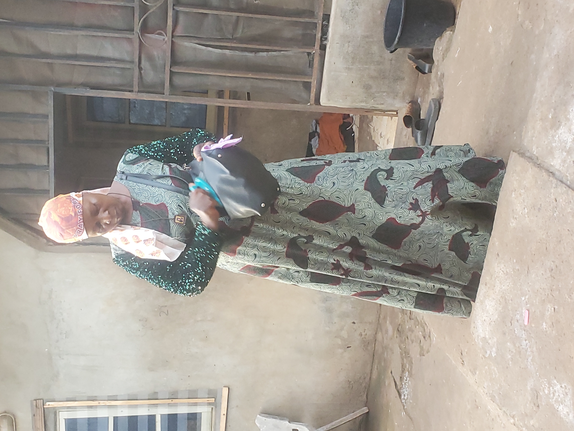 A woman stands in what appears to be an outdoor courtyard or enclosed area. She is wearing a patterned dress and head covering, and she carries a black handbag. The environment is rustic, with concrete walls, a simple window, and basic metal structures overhead. There are items such as a bucket and what seems to be footwear nearby. The location is Ukwu-Abwa, Nigeria. There is not any activity or interaction evident beyond the woman's presence.