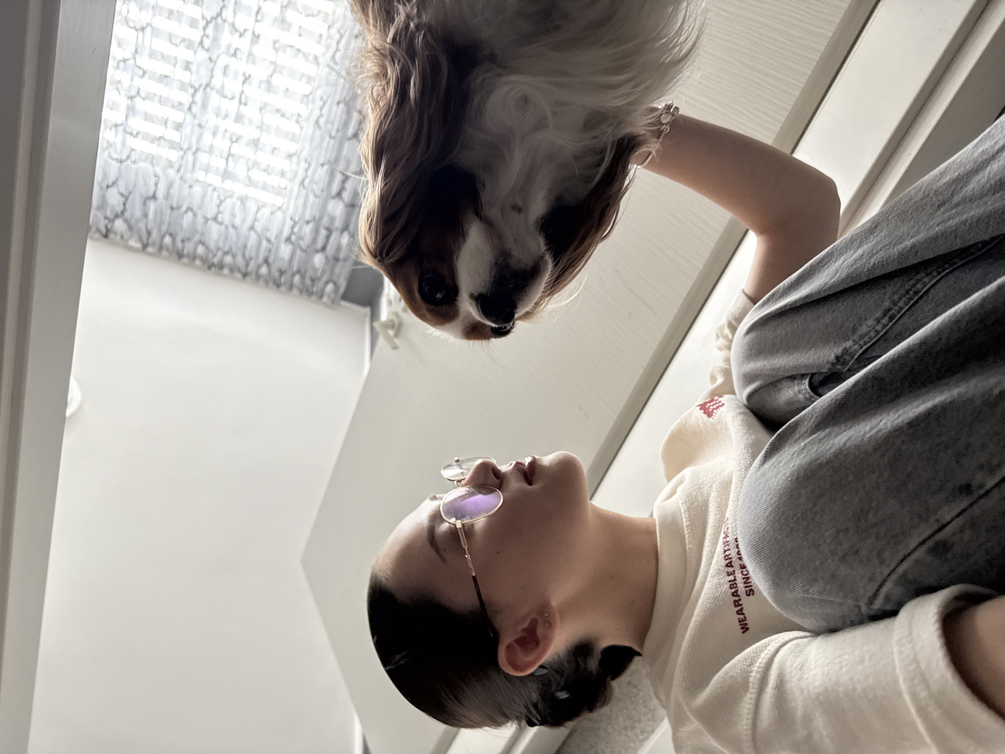In a well-lit interior, a young woman with dark hair pulled back and wearing glasses crouches down to interact with a Cavalier King Charles Spaniel. The woman is dressed in a cream-colored sweatshirt with text on it and light-wash jeans. Her left hand is visible, resting on the dog. The dog, facing upwards, has a brown and white coat and is looking at the woman. The background features a white wall and a window with patterned curtains. The overall impression is one of affection and connection between the woman and her dog.