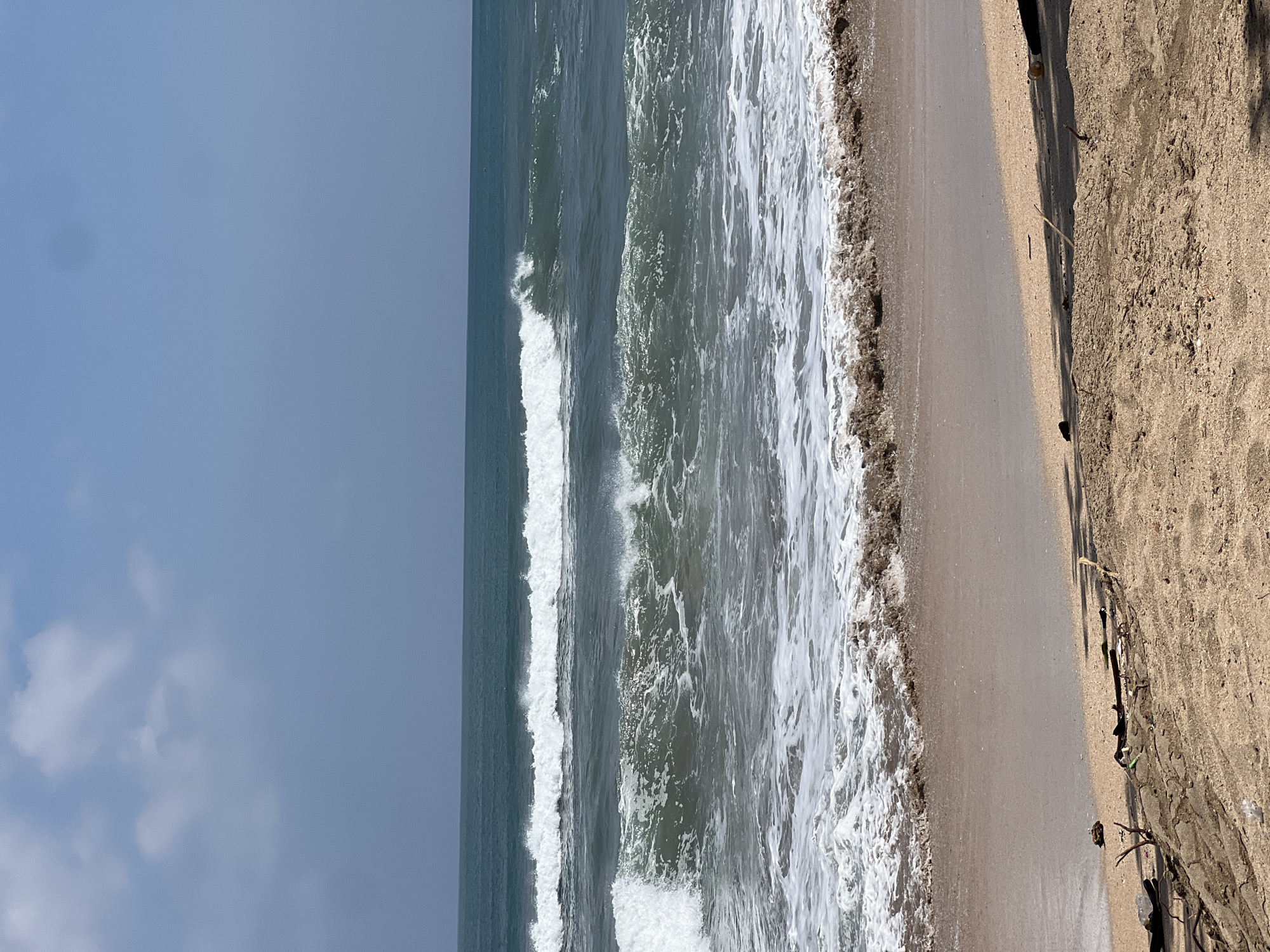 The image shows a beach scene in Kuta, Indonesia. The foreground is dominated by sand, with some driftwood and debris scattered across it. In the midground, the ocean is visible with gentle waves rolling towards the shore, creating white foam as they break. The water appears a deep blue-green. The background consists of a clear sky with a few wispy clouds. The overall impression is one of a tranquil, natural coastal environment.