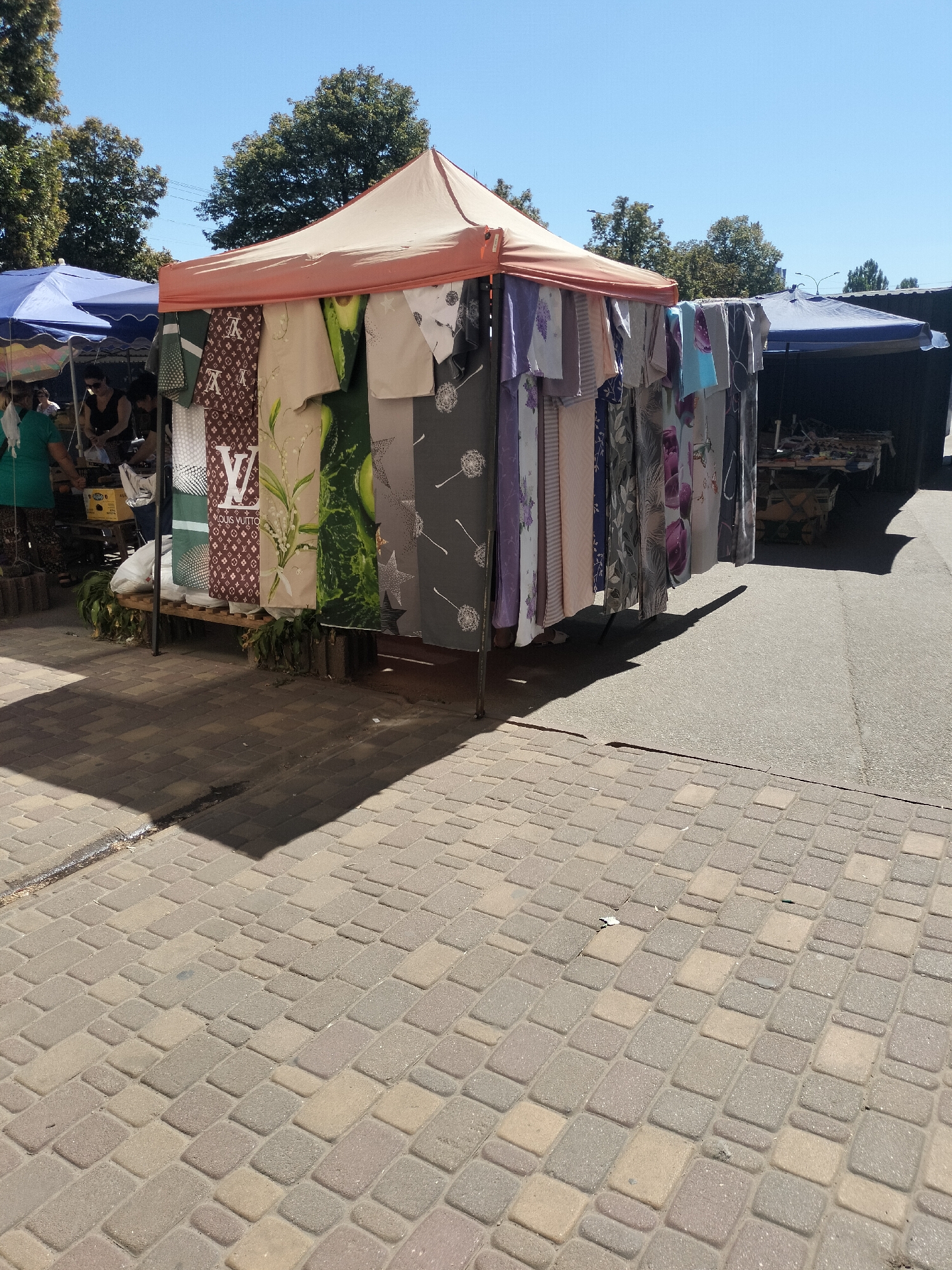 A street market scene on a sunny day in Chernihiv, Ukraine, with stalls selling textiles. In the foreground, patterned paving stones cast long shadows. A temporary stall with an orange canopy displays various fabrics, including one with a repeating "Louis Vuitton" pattern and another with an avocado design. To the left, another stall with a blue umbrella is partially visible, with people attending to merchandise. Further in the background, more market stalls and trees line the street, under a clear blue sky.