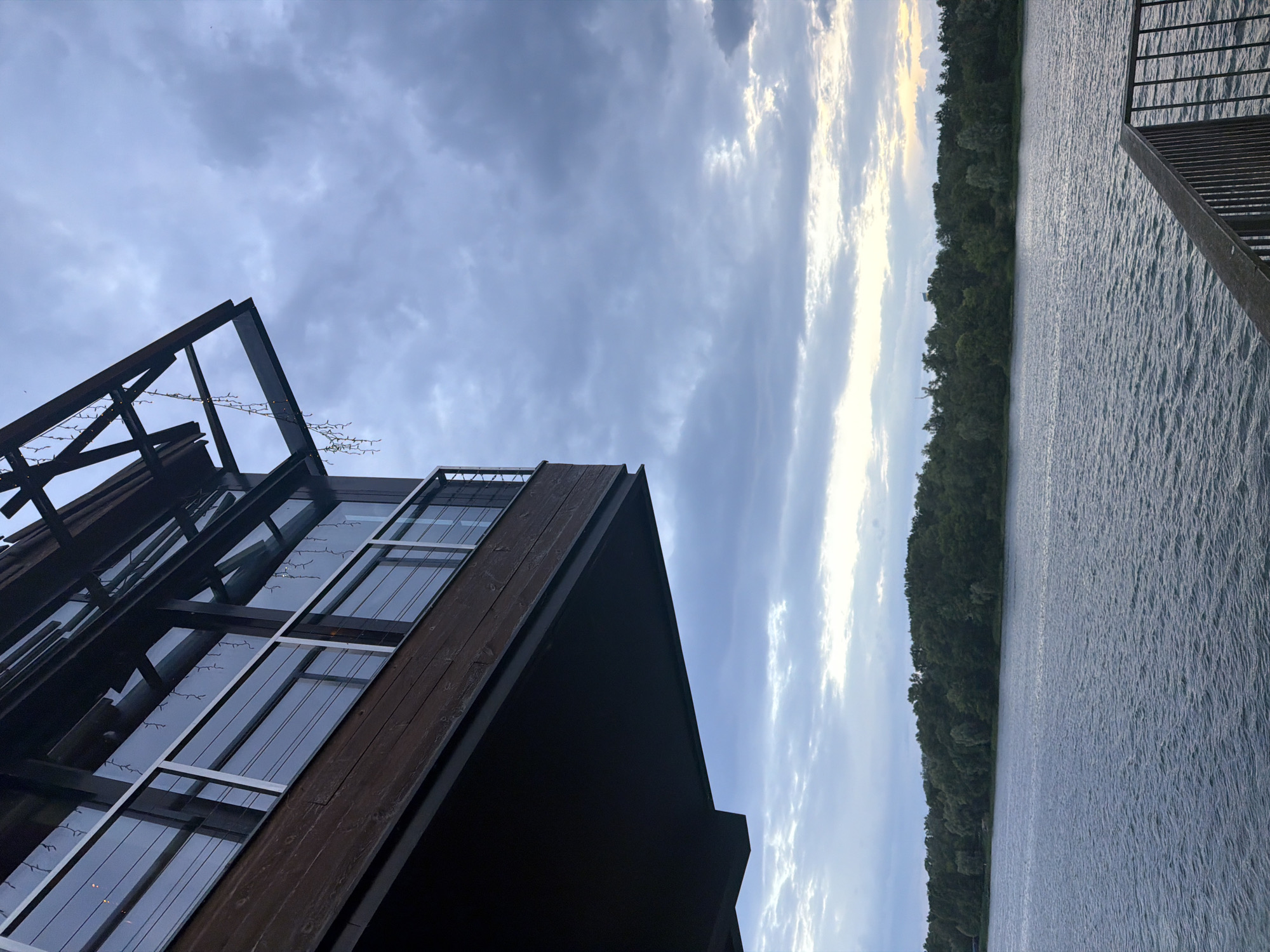 This is an image of a modern wooden and glass building with a dark facade situated on the shore of a body of water. The building has large windows that reflect the sky. A metal railing is visible on the right side of the image, suggesting a deck or walkway. The water has small ripples, indicating a gentle breeze. A dense line of trees forms the background, with a sliver of light sky visible above them, suggesting it might be dusk or dawn. The clouds are a mix of grey and white, with some areas catching the light. The overall mood is serene and peaceful. The image is oriented vertically. There is no visible text.