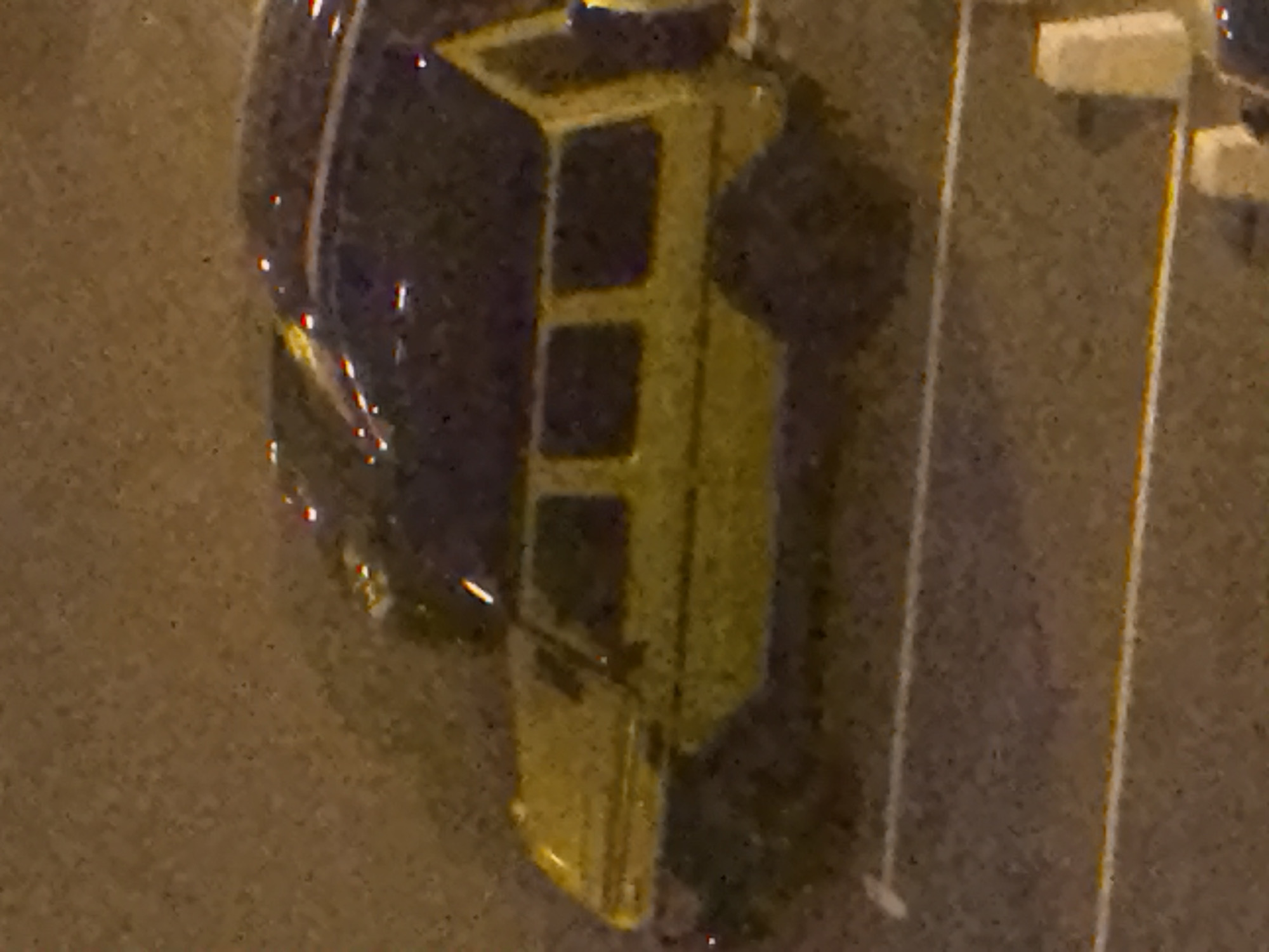 A yellow vehicle with a boxy shape, resembling a Mercedes-Benz G-Wagen, is parked on a dark asphalt surface. The vehicle has tinted windows and appears to be a four-door model. It is positioned on a slight incline, with the front of the vehicle angled slightly upwards. In the background, another dark-colored vehicle is partially visible, its shape obscured by the lighting. The scene appears to be an outdoor parking lot, possibly in a city environment, given the presence of what seem to be painted lane markings on the asphalt. The lighting suggests it is nighttime or dusk. There are no people visible in the image. There is no information to confirm the location as Moscow, Russia.