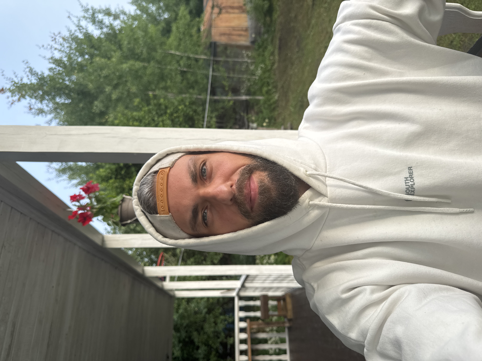 A mid-shot, selfie-style photograph captures a man with a beard and grey-streaked hair, wearing a white hoodie and a beige baseball cap, with the hood pulled up. He is positioned on what appears to be a porch or veranda, with white railings visible to his left. In the background, lush green trees and a rustic wooden shed are discernible under a clear blue sky. The overall setting suggests a rural or suburban environment, possibly in or around the city of Kuti, Russia, given the architecture and natural surroundings. The man is looking directly at the camera with a neutral expression. The lighting suggests it is daytime, possibly morning or late afternoon due to the soft light.