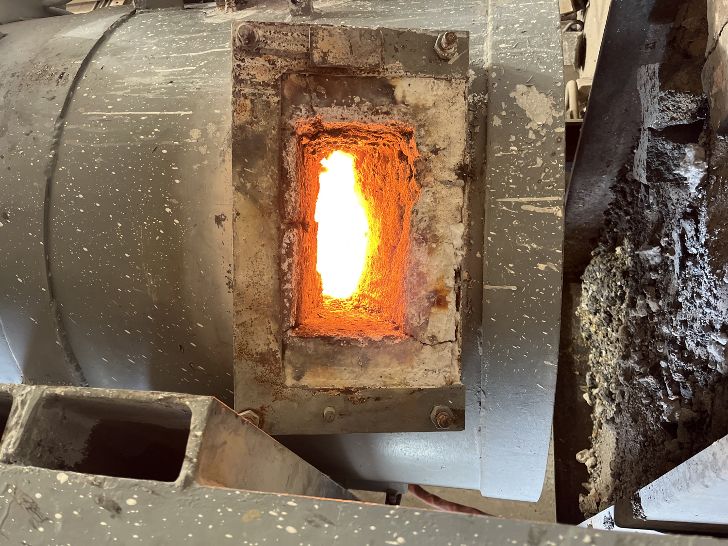 The image depicts an industrial scene centered on an active furnace or kiln, likely operating at high temperatures within an indoor facility. A prominent rectangular opening in the center reveals a bright, intense orange-yellow flame or glowing heat emanating from inside. The interior of this port appears to be lined with worn refractory material, indicative of prolonged exposure to extreme heat.
To the left, a large, curved grey metal surface is visible, heavily speckled with white splatters, suggesting it's part of a cylindrical component such as a rotary kiln or similar industrial heating apparatus. This section shows visible seams and fasteners. To the right of the fiery opening, a pile of dark, granular material, possibly solidified slag, ash, or processed material, is accumulated on the ground or a platform.
In the bottom left foreground, a robust, hollow grey metal structure, potentially a support beam or part of the machinery's frame, is partially visible. The entire environment appears utilitarian and industrial, with surfaces showing wear, dust, and spatters consistent with heavy use in a manufacturing or processing plant. No individuals are present in the image, and there is no visible text. The intense glow from the furnace signifies an ongoing, high-temperature industrial process at this location in Saint-Rémy-lès-Chevreuse, France.