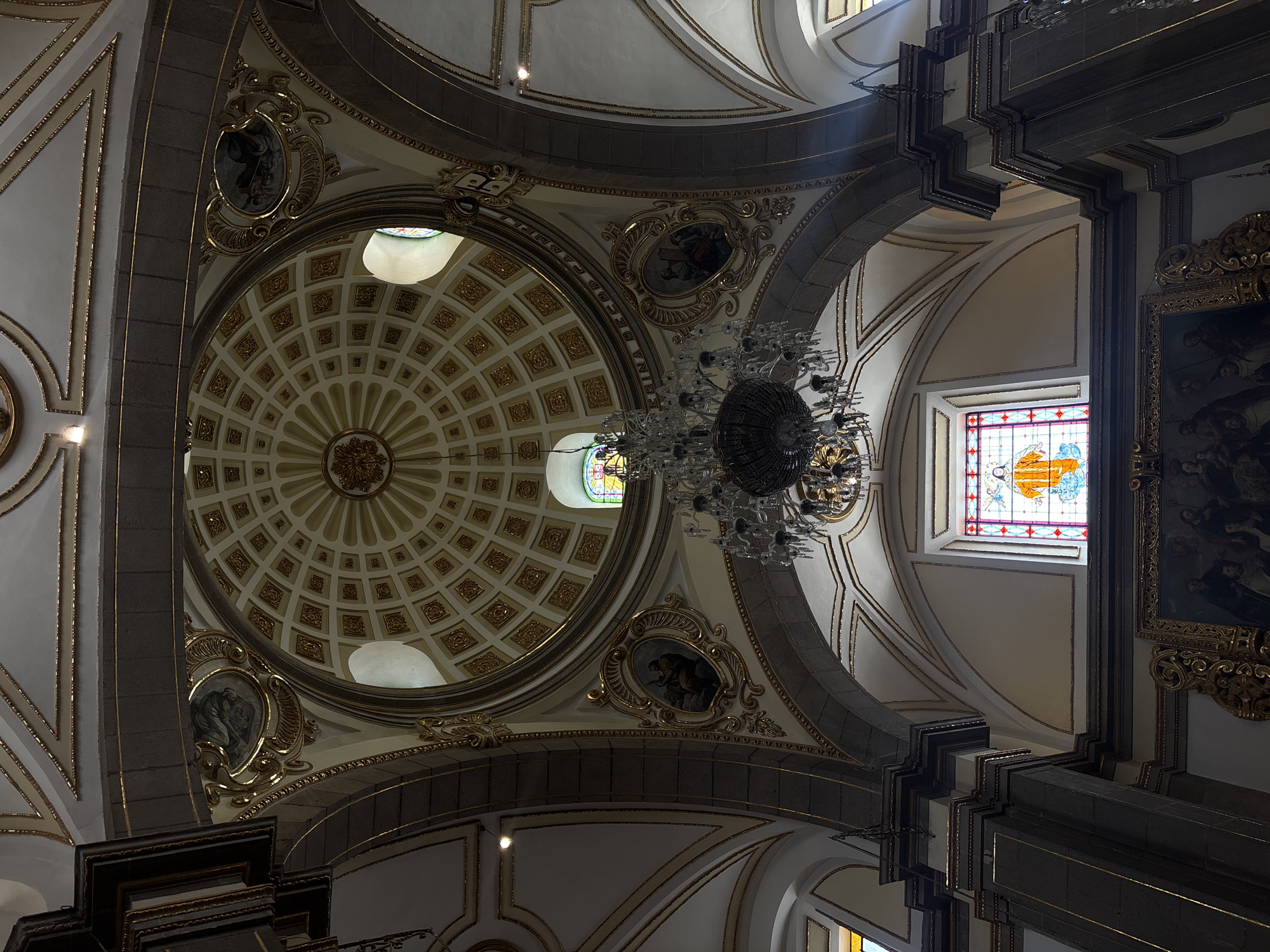 The image captures the interior of a church, likely a cathedral, in Puebla, Mexico, showcasing the architectural details of the dome and supporting structure. The dome is adorned with intricate gold and white patterns, radiating from a central medallion. Around the base of the dome are several oval-shaped frames containing religious paintings. A large chandelier hangs prominently in the foreground, partially obscuring the view. The architecture includes arches, pillars, and a stained-glass window with a colorful design. A framed painting hangs on one of the structural elements, depicting a scene with multiple figures. The style of the architecture and decoration suggests a Baroque or Neoclassical influence.