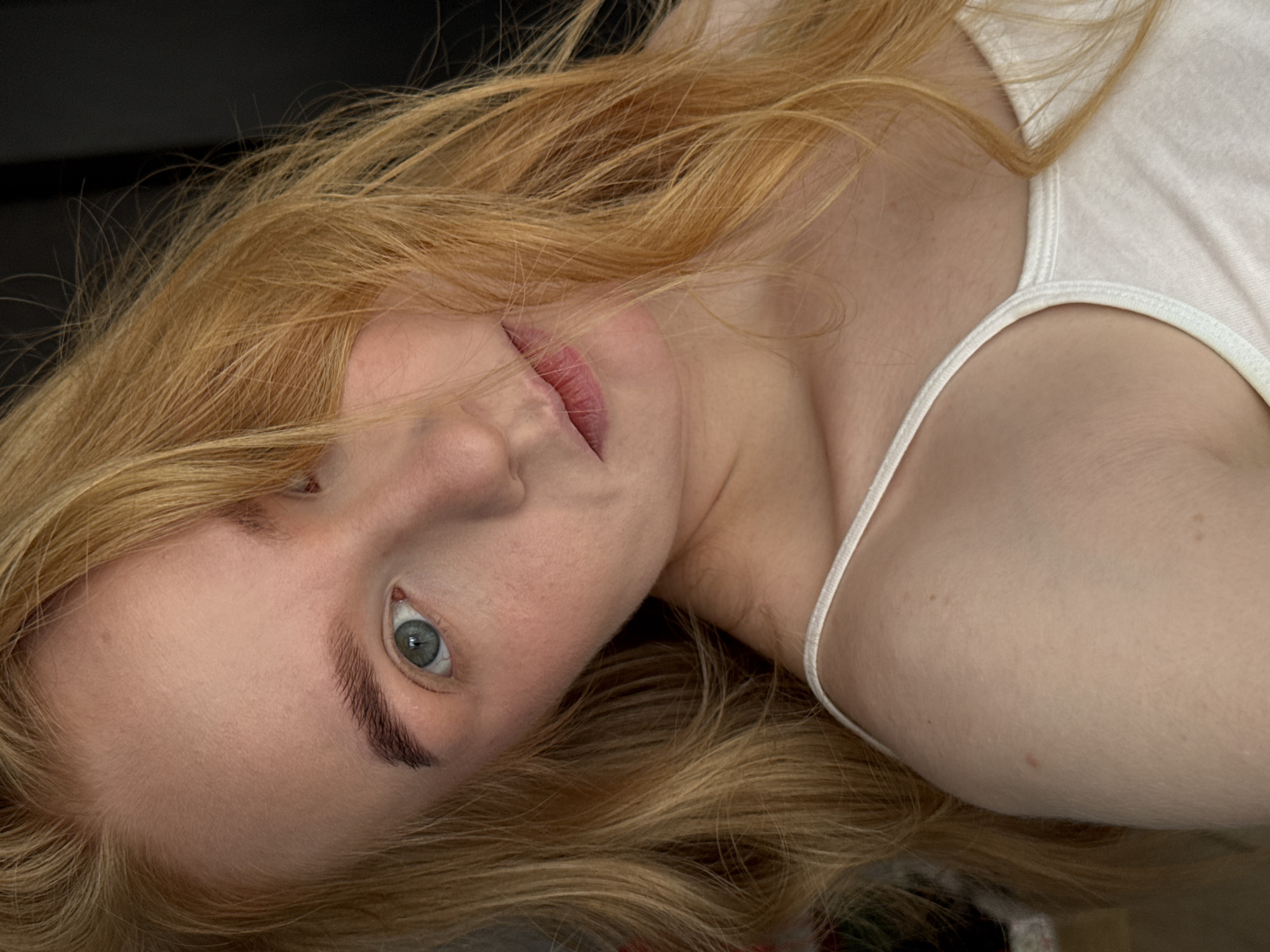 A pale-skinned young woman with long strawberry blonde hair is angled in the frame, facing the camera. Her head is tilted to the right. Her expression is neutral. She has light eyebrows and visible blue-gray eyes. Her lips are slightly parted. She is wearing a white tank top. The background is dark and mostly out of focus. No identifying landmarks or elements suggest the location of Chaikovsky, Russia.