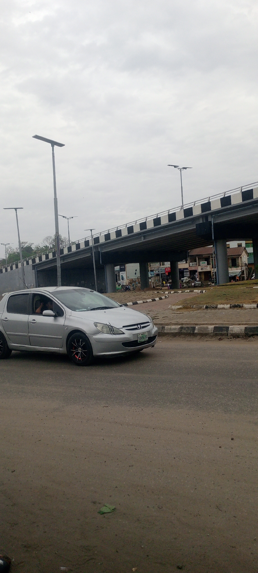 A silver Peugeot 307 hatchback, registration number SAN-961-LA, is traveling on a paved road in Adankolo, Nigeria. The car has black rims with red accents and sustained minor body damage. The road shows signs of wear and tear. An overpass runs above the road, supported by concrete pillars. A row of street lights lines the overpass. Businesses and residential buildings are visible in the background below the overpass. The sky is overcast.