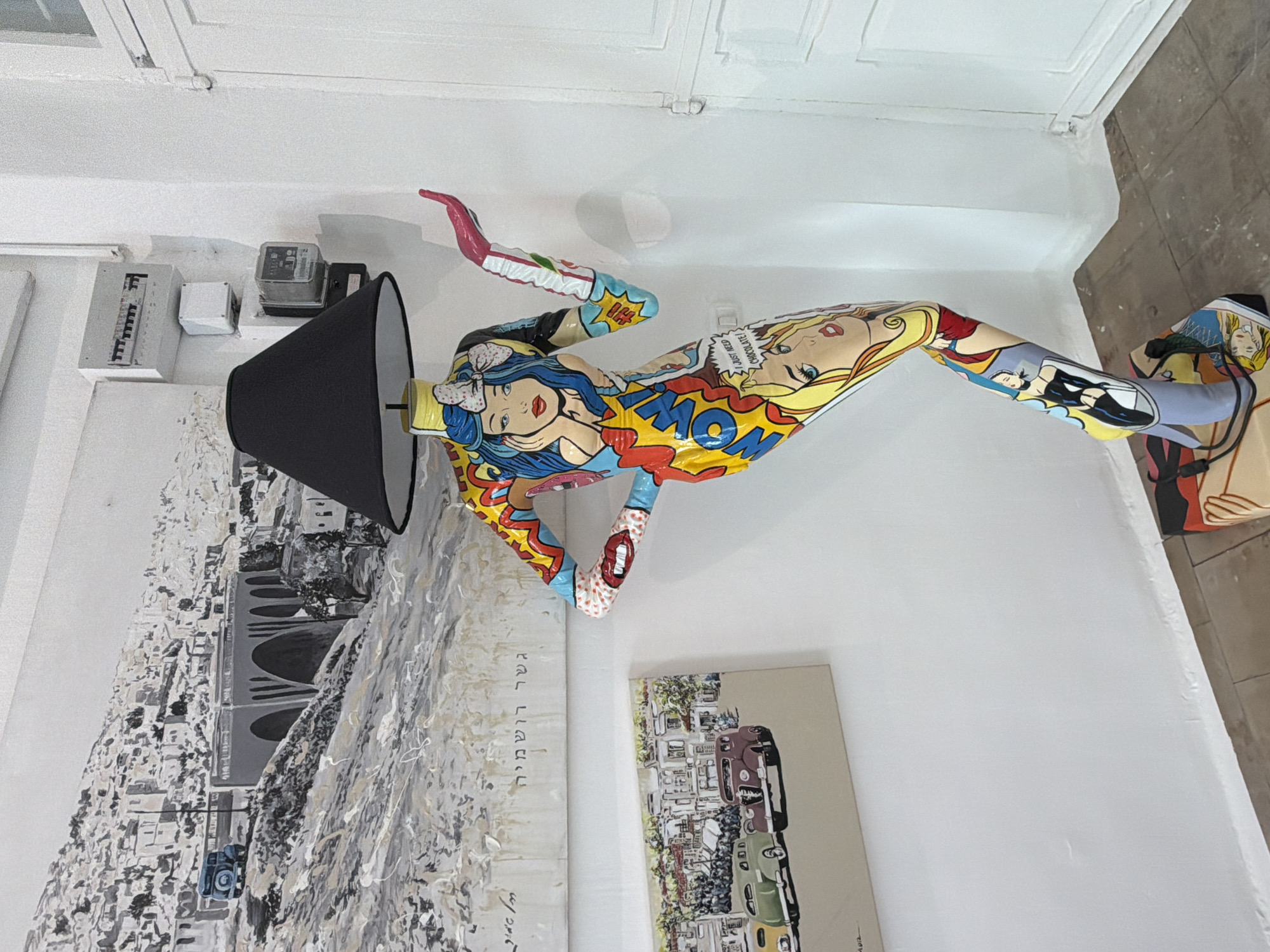 The image shows an indoor scene, possibly a gallery or a living space, featuring an eclectic mix of art and objects. Dominating the scene is a pop-art style sculpture of a woman in a reclining position, adorned with bright colors and comic-book motifs. The sculpture is placed next to a black lamp.
On the wall behind the sculpture is a large black and white painting depicting what appears to be an urban landscape featuring a bridge. To the right of this painting is a smaller color painting of cars and city streets.
In the background, there is evidence of standard electrical fixtures and a light switch. The floor and walls are neutral in color, and the overall lighting appears to be bright. The scene evokes a sense of artistic expression and cultural flair. I cannot confirm the specific location in Tel-Aviv, Israel.