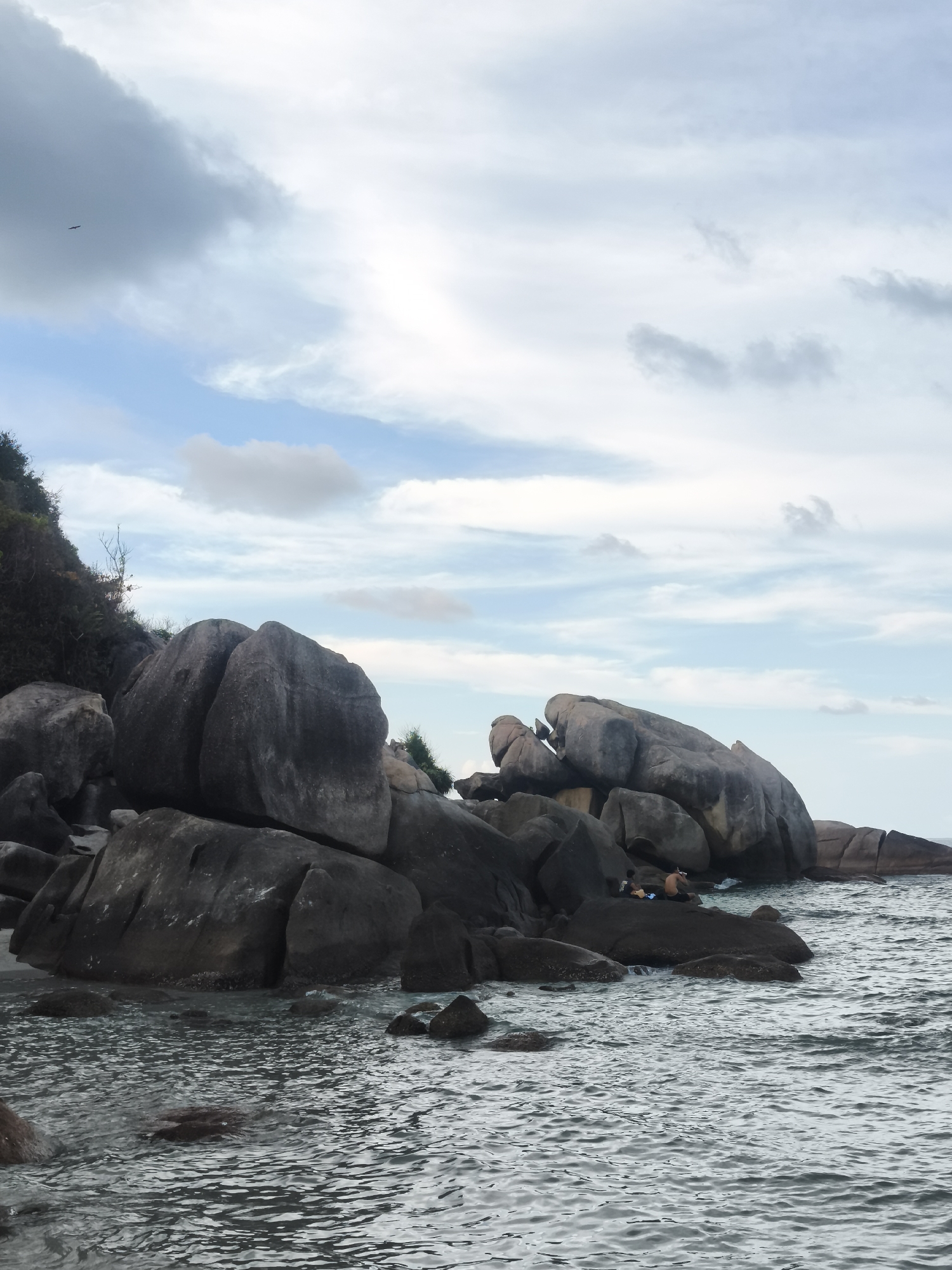 The image depicts a rocky shoreline in Baan Chaweng Noi, Thailand. Large, dark grey granite boulders dominate the scene, extending into the water. The water is grey with visible ripples and small waves. Two individuals are present on one of the larger rocks. The sky is partially cloudy with a mix of dark grey and light blue patches. A small bird is visible in the upper left portion of the frame. Trees and other greenery are visible atop the rocks.