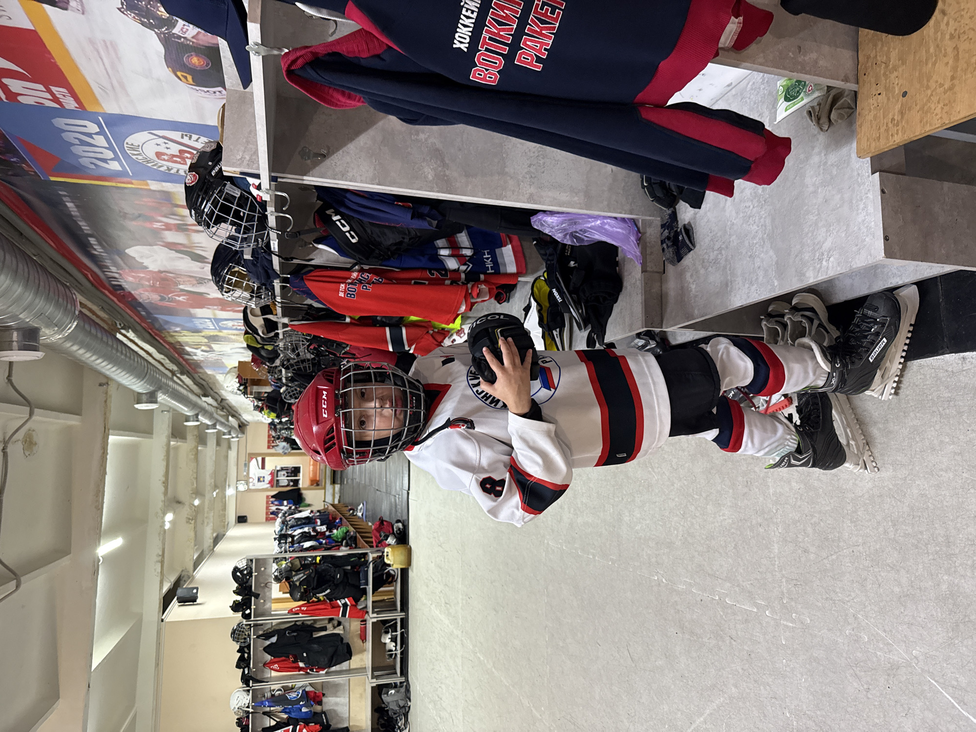 A young person in full hockey gear stands in a locker room, holding a hockey puck. The person is wearing a red helmet, a white and black jersey with the number 8, black hockey pants, and skates. The locker room is equipped with shelves and hooks, holding various hockey equipment and clothing. A large banner with "2020" and Russian text is visible in the background. The floor is made of ice, indicating the location is an ice rink. The text on the jersey and the banner suggest the location is Votkinsk, Russia.