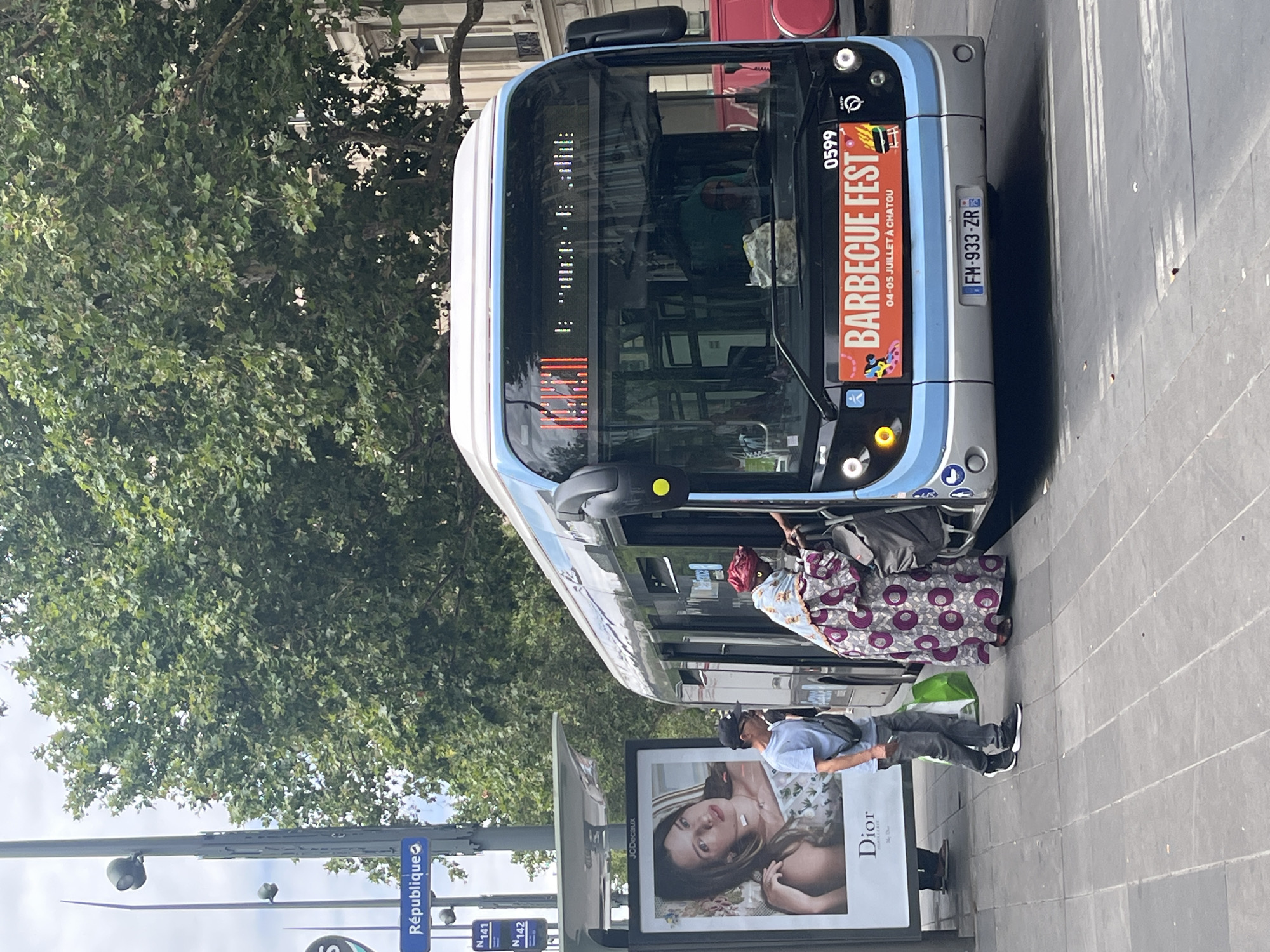 This media file shows a street scene in Paris, France, with a bus as the central object. Two people are visible near the bus entrance. One, wearing a headscarf and a patterned dress, appears to be boarding or disembarking. The second, a man in casual clothing, stands nearby, looking towards the bus.
The bus is a light blue and grey public transport vehicle, displaying "BARBECUE FEST 04-05 JUILLET À CHATOU" on its front, suggesting an event promotion. The license plate reads "FM-933-ZR". The bus also has an electronic display above the windshield showing a line of text that is partially obscured.
In the background, a lush green tree provides shade, and parts of buildings can be seen. In the foreground, a large advertisement for Dior is visible, featuring a woman's face. Street signs with "République", "N141", and "N142" indicate a public transportation hub. The ground is paved with grey tiles.
The scene suggests a daytime setting, likely a warm day given the foliage and attire of the people. The overall atmosphere appears to be that of everyday city life with a hint of an upcoming event.
