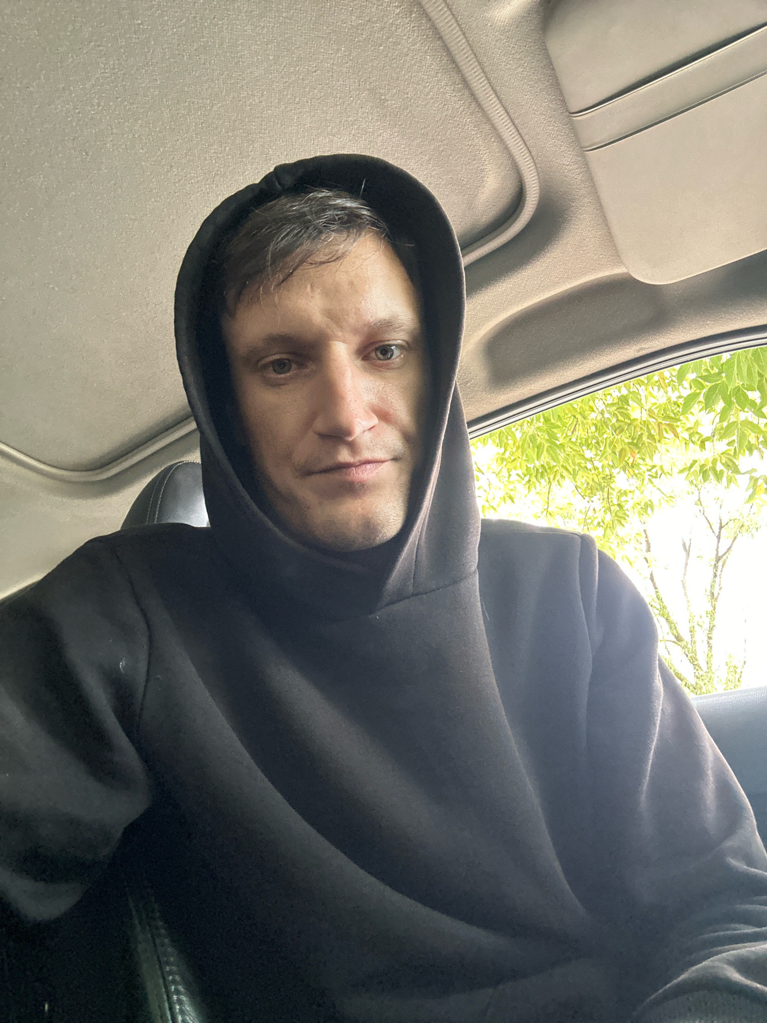 A man is seated in a car, wearing a black hooded sweatshirt. His face is turned towards the camera, and he has short, dark hair. The interior of the car, including the ceiling and a sun visor, is visible. Through the car window, green foliage and trees are visible, indicating an outdoor setting. The exact location in Makiv, Ukraine, cannot be determined from the image alone.